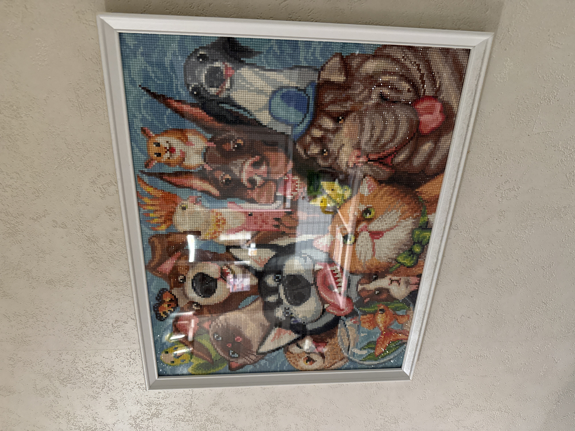 The image shows a framed diamond painting of many animals, predominantly dogs, on a textured wall. The painting is horizontally oriented and is hung at eye level. The animals are depicted in a cartoonish style with a blue background. Some of the animals visible include a husky with its tongue out, a brown dog with floppy ears, a black and white dog with a blue hat, a cat with pink ears, and other smaller animals like a hamster and guinea pig. There is also a butterfly in the upper left corner and a goldfish in a bowl at the bottom left. The overall impression is a cheerful and colorful artwork. The wall appears to be painted with a light-colored, textured finish.