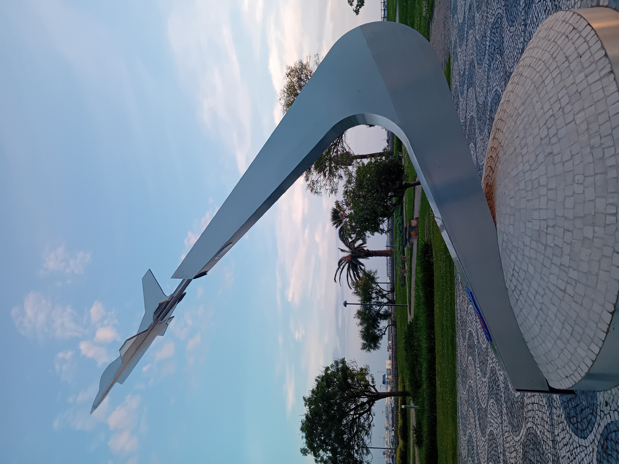 The image shows a monument in Figueira da Foz, Portugal, likely the "Air Force Tribute" (Monumento Homenagem à Força Aérea). The monument consists of a stylized representation of an aircraft wing intersecting with a circular mosaic platform. The "wing" is a large, curved metal structure. The ground around the base of the monument is covered in patterned cobblestones. A grassy park area with trees, including a palm tree, stretches behind the monument. In the background, a body of water and possibly some buildings are visible on the horizon. The sky is light blue with scattered clouds.