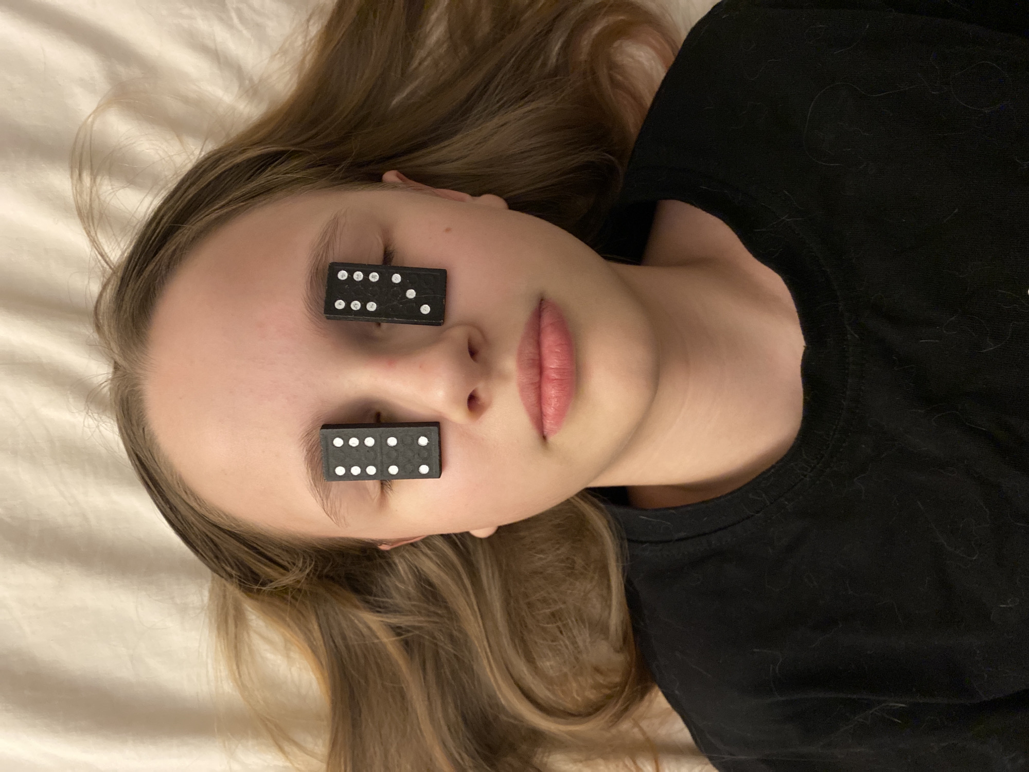 A person lies on their back on a light-colored fabric, possibly a bed sheet. The person has light skin, light brown hair with blonde highlights, and their eyes are covered by two black dominoes, each displaying six white dots. The person is wearing a black t-shirt. There is no evidence to suggest the location is Yekaterinburg, Russia.