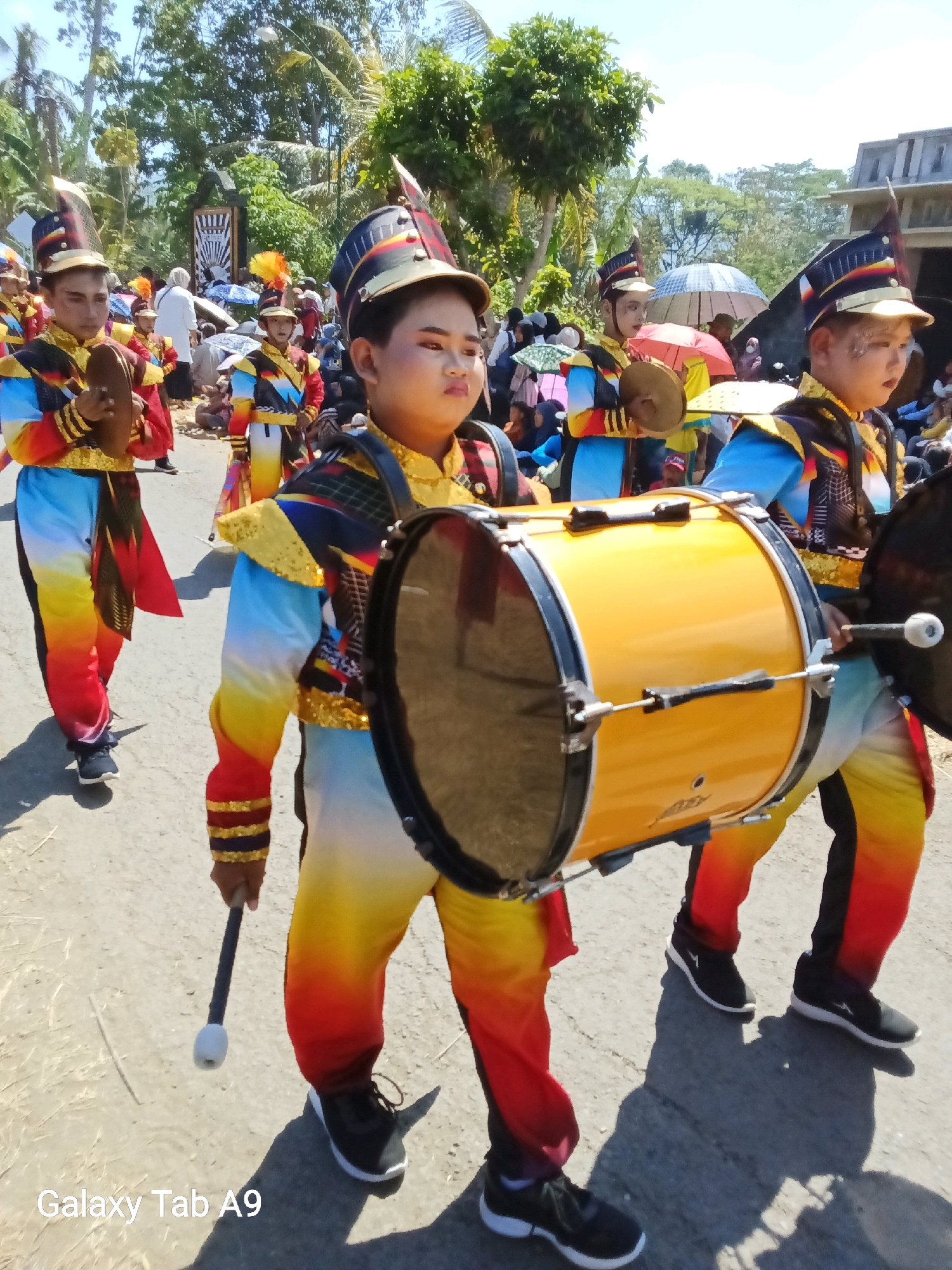 A group of children in brightly colored marching band uniforms are parading down a paved road. The children are wearing tall hats with plumes, jackets with gold trim, and pants that transition from yellow to orange to red. Some are carrying drums, while others have smaller percussion instruments. In the background, a crowd of spectators watches the parade under a bright, sunny sky. Trees and buildings are visible along the sides of the road. The scene appears to be a festive community event in Ngadirenggo, Indonesia.