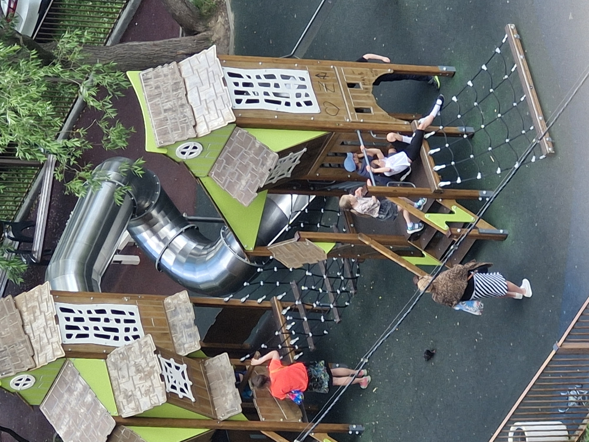 The image depicts a playground scene in the city of Ерино, Russia, dominated by a multi-level wooden play structure with green accents and a large silver slide. Several children and one adult are present. A young person in an orange shirt and patterned skirt stands on a platform of the play structure, facing away from the camera, holding a multi-colored bag. Another adult, wearing a patterned shawl and a striped skirt, stands on the green rubberized ground, also with their back to the camera, holding a shopping bag.

Within the play structure, multiple children are visible. Two children are on a lower level, one leaning on a horizontal bar and the other sitting on a platform. Another child, wearing a grey top and dark skirt, stands to the left, looking towards the play structure. To the right of the structure, a child in a white t-shirt and black pants is hanging from a horizontal bar with their legs extended. A young child in a blue cap is sitting on a platform. A person's legs are visible on the far right side, wearing dark pants and bright green sneakers, suggesting they are standing or interacting with the structure. The playground surface is a dark green, textured rubber. A large tree with green foliage is situated behind the play structure. A metal railing and a trash bin are visible in the lower left portion of the image. The overall environment suggests a park or recreational area.