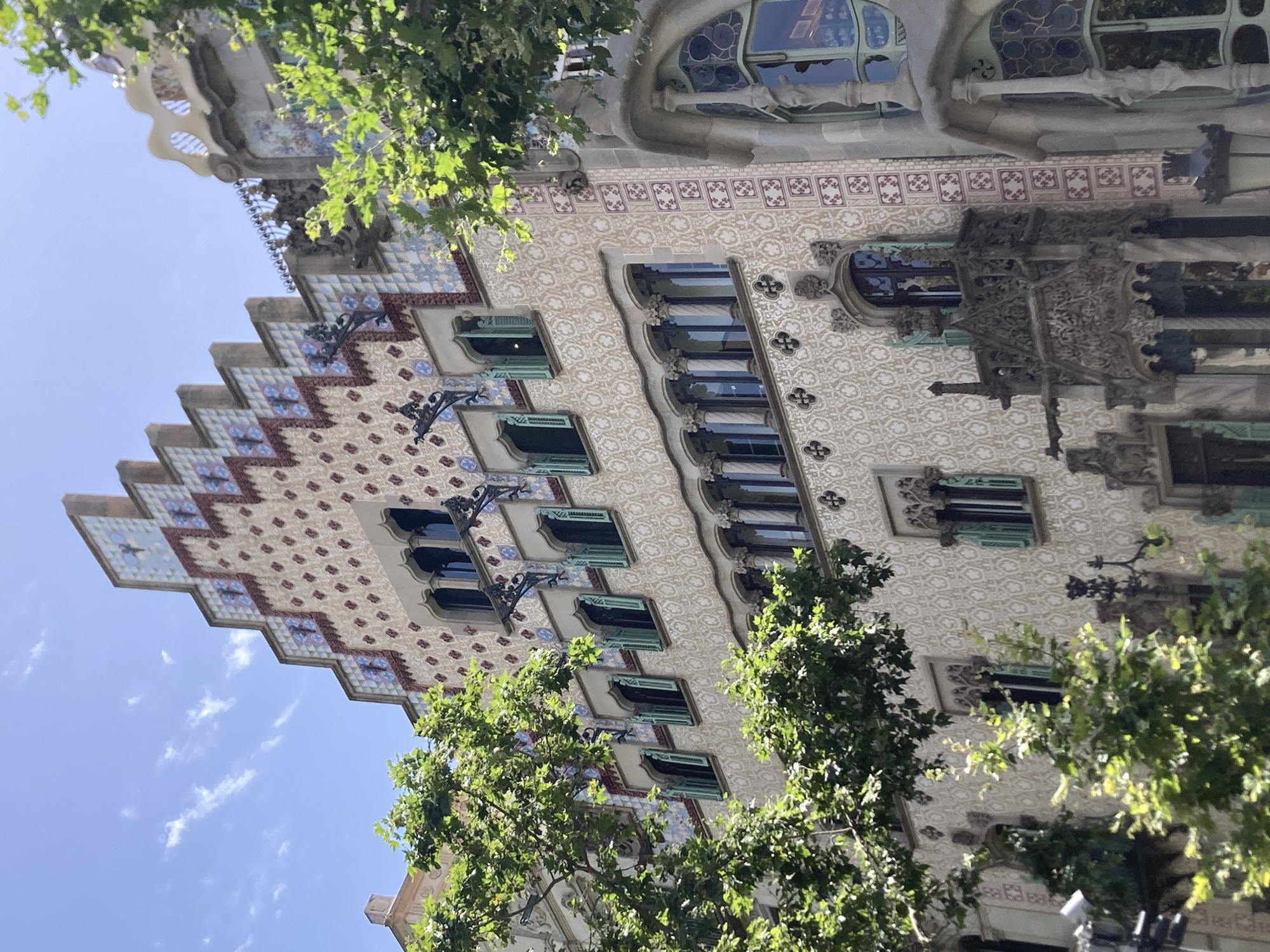 The image shows a detailed view of the Casa Batlló, a renowned building located on Passeig de Gràcia in the Eixample district of Barcelona, Spain. The facade is characterized by its organic, undulating forms and vibrant ornamentation. The upper part of the building features a stepped roofline adorned with colorful ceramic tiles in shades of blue, white, and red, arranged in a zigzag pattern. Below the roof, a series of windows with arched frames are visible, many fitted with light green shutters. The main body of the facade is covered in a textured mosaic of small, light-colored tiles, creating a shimmering effect. The building's design incorporates decorative elements such as wrought-iron balconies with intricate designs, sculpted gargoyles, and elaborate window frames. Green trees with dense foliage frame the building from the left and right, partially obscuring the lower sections and the street level. The sky above is a clear blue with scattered white clouds. The architectural style is distinctly Modernista, with a sense of playful fantasy and organic inspiration. There are no people or animals visible in the frame.