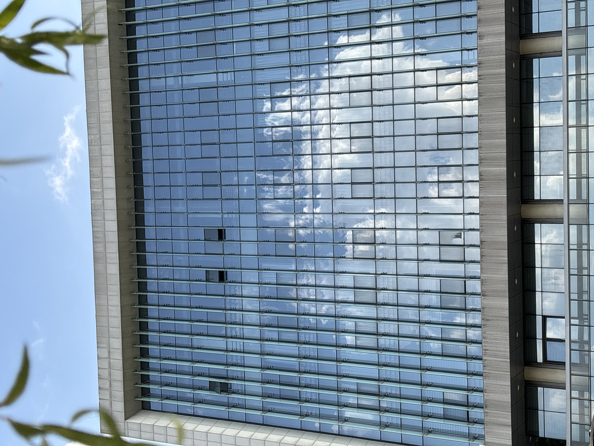 A low-angle, full-frame shot captures the facade of a modern building with a glass curtain wall, reflecting a blue sky with white clouds. The building has a concrete upper structure with a row of vertical louvers above the glass. The glass facade is divided into a grid of rectangular panes, with some windows slightly ajar. The lower portion of the image reveals another level of the building with a different window arrangement and a pedestrian walkway with a glass railing. Foliage is visible in the upper left and right corners, partially obscuring the sky. The image was taken in Jiangjin, China.