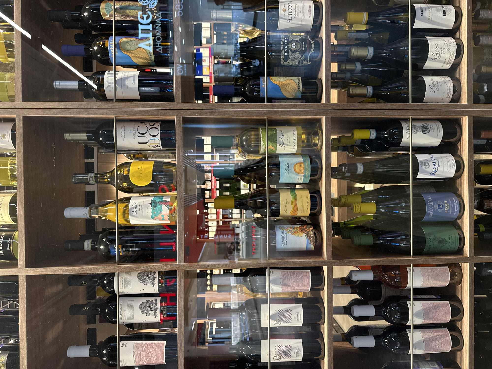 The image is a medium shot of a wine display shelf in a store. The shelf is divided into two vertical sections by a wooden divider. The shelves are made of wood and are filled with various bottles of wine.
In the left section of the shelf, there are several rows of wine bottles. The top row shows bottles with white, red, and yellow labels. The second row from the top features several dark-colored wine bottles with white and red labels. The third row contains a mix of dark and lighter-colored wine bottles, some with white labels and others with colorful designs. The bottom row displays more dark-colored wine bottles with white labels.
The right section of the shelf also contains rows of wine bottles. The top row shows dark-colored bottles with white and blue labels. The second row features dark-colored bottles with white and colorful labels, including a bottle with an illustration of a person on it. The third row has dark-colored bottles with white labels and some with intricate designs. The bottom row displays dark-colored wine bottles with white labels and some with colorful graphics.
The bottles are arranged in an organized manner, with some rows appearing fuller than others. The lighting in the store is visible as a bright white strip along the top edge of the right section, suggesting overhead lighting. Reflections on the glass in front of the bottles indicate a display case or a similar protective barrier. The overall impression is a well-stocked wine cellar or retail display.