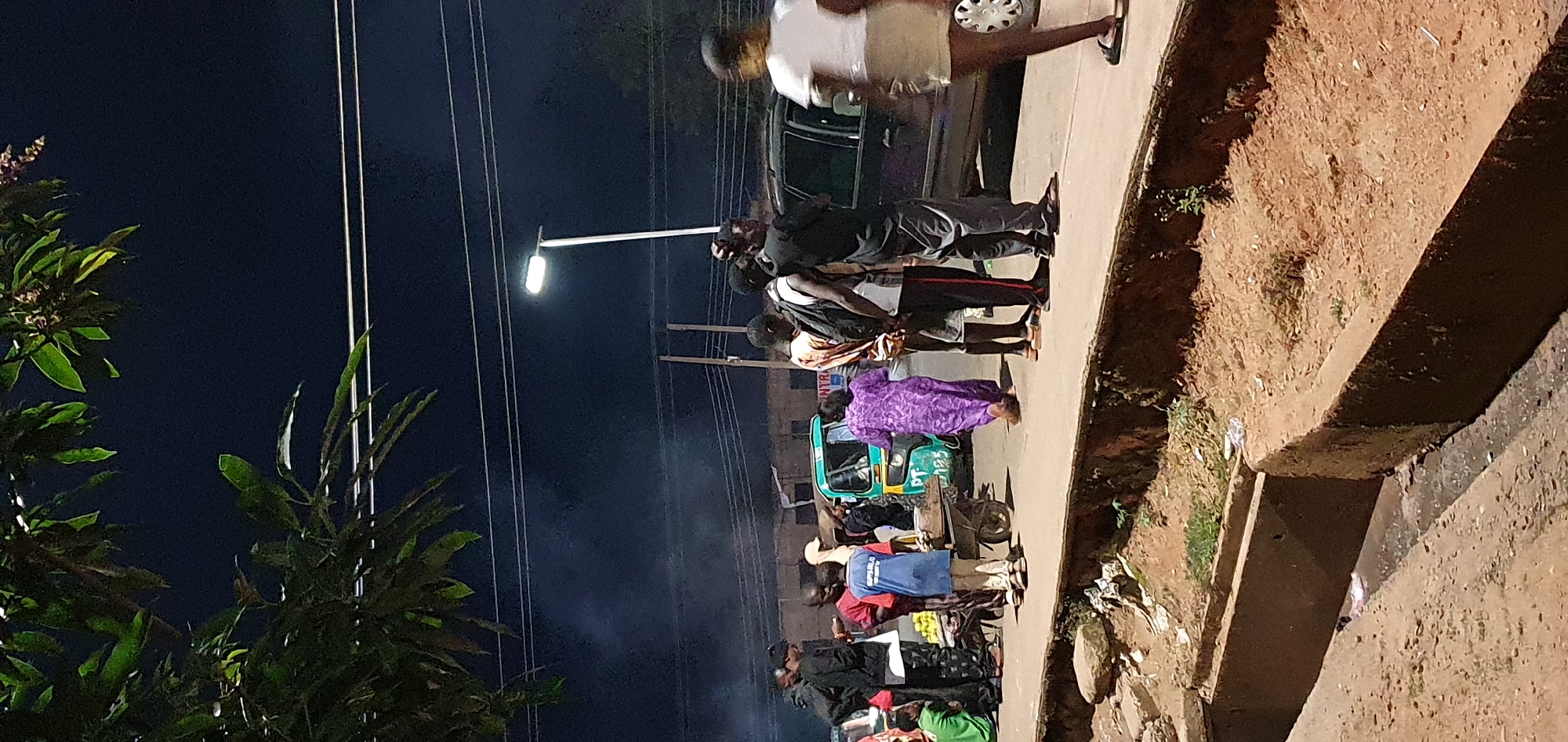 The image depicts a nighttime street scene in Bukuru, Nigeria, with a group of people gathered near a road. In the foreground, a drainage ditch is visible, partially filled with murky water and debris. The midground features a paved road where several individuals are standing. A bright street lamp illuminates the area. A teal and yellow auto-rickshaw is parked on the side of the road, with a cart laden with items next to it. A woman in a purple patterned dress is walking away from the rickshaw. Several men and boys are standing around, some with backpacks. One boy in a blue shirt appears to be interacting with the cart. In the background, a dark-colored SUV is parked, and building facades are dimly visible. The sky is dark, with some faint wisps of smoke or mist present. A large tree with green leaves hangs into the frame from the top left corner.