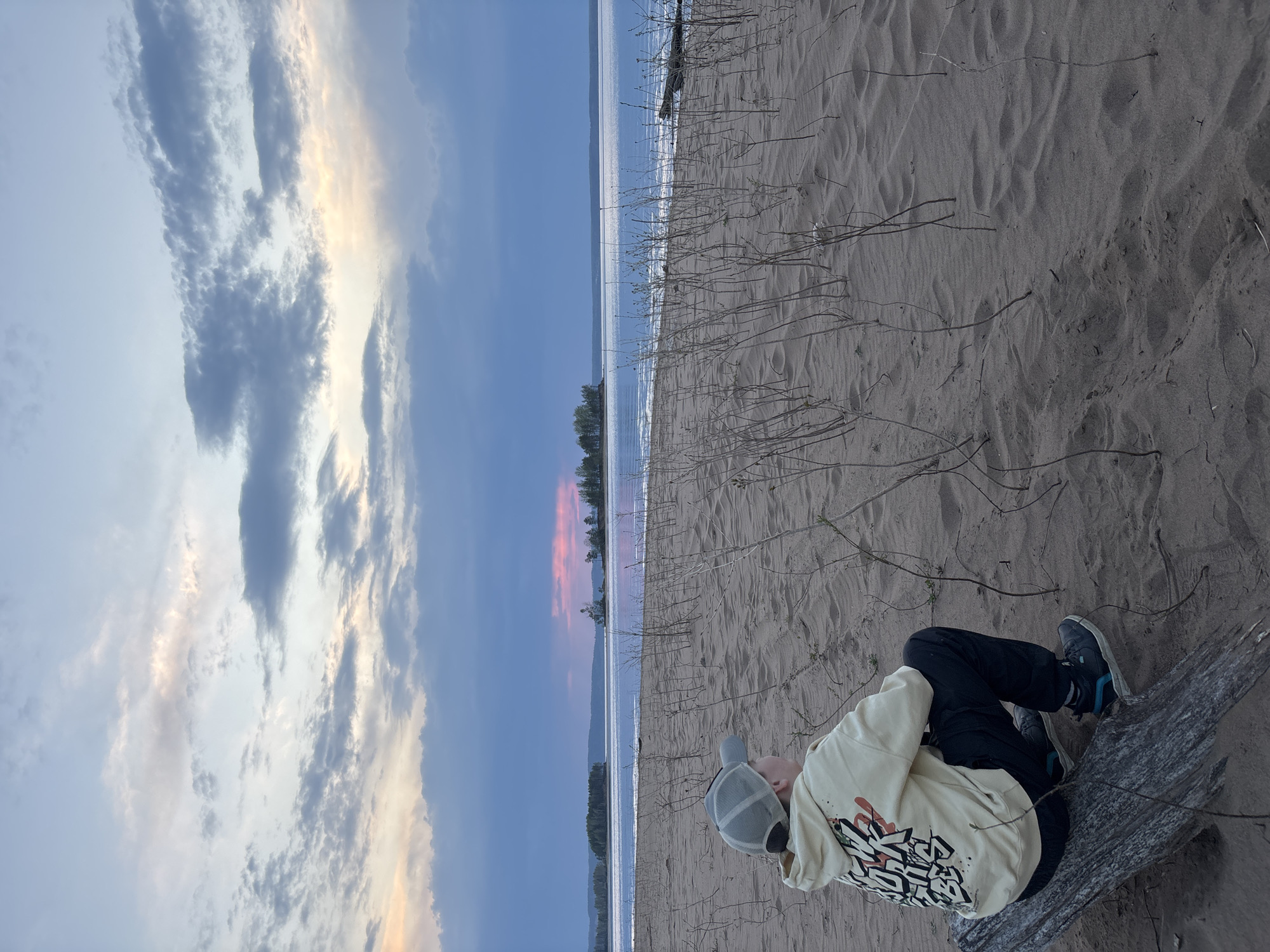 A horizontal outdoor shot presents a person on a sandy shore at what appears to be dusk.
The sky is a mix of light blue and clouds tinged with orange and pink hues, suggesting a sunset or sunrise. The body of water adjacent to the beach reflects the colors of the sky, creating a mirrored effect.
The sandy beach is the primary foreground element, marked with small patches of dry vegetation and the texture of footprints. A person is resting on a weathered piece of driftwood. They are wearing dark pants, sneakers with visible blue accents, and a light-colored hoodie. The hoodie is adorned with a graphic design on the back. The person is also wearing a baseball cap.
The environment is a beach, possibly in the city of Vekoshinka, Russia. The image captures a moment of solitude by the water.