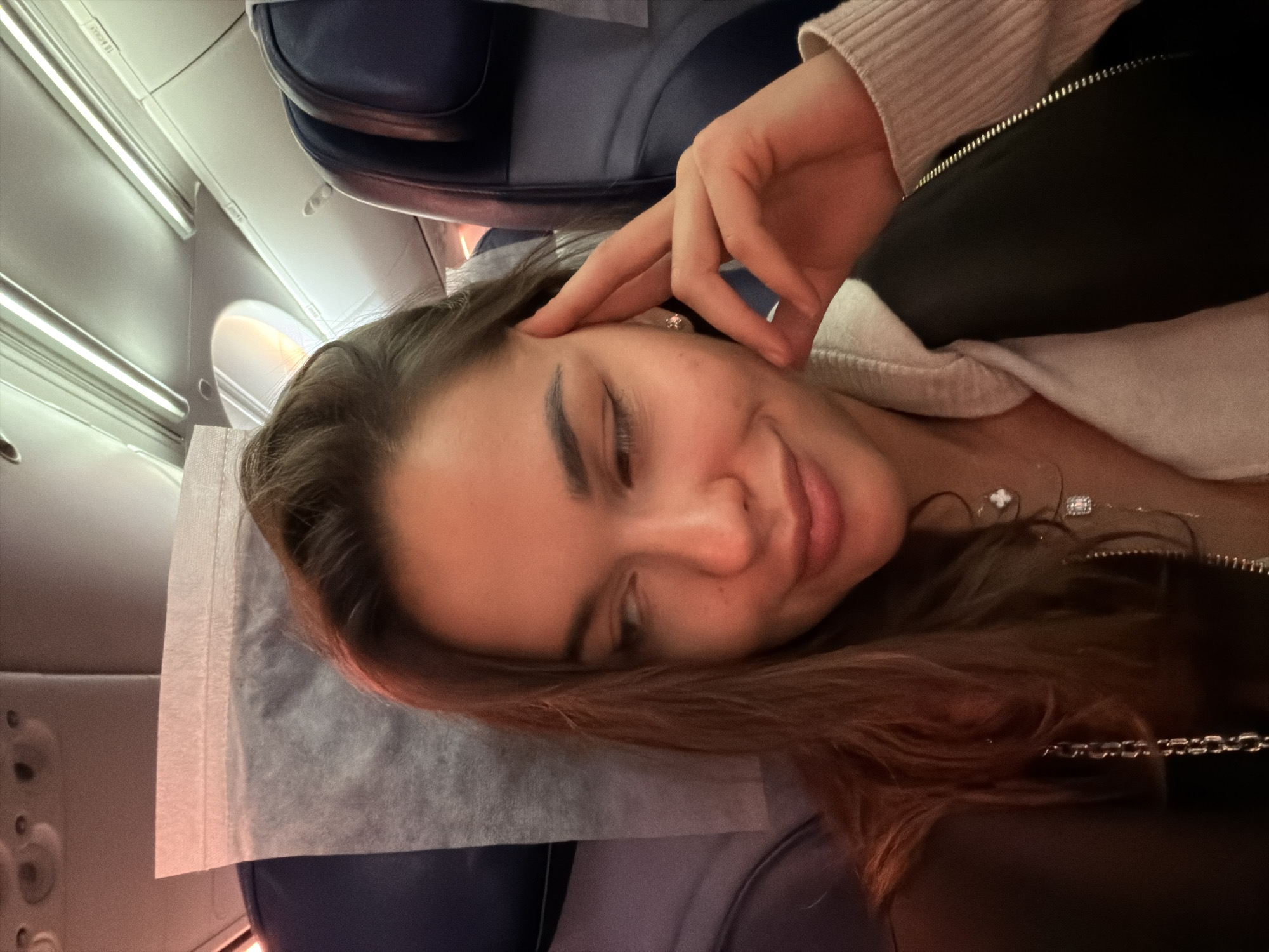 A woman is in what appears to be an airplane cabin. She is reclining with her head on a pillow, smiling slightly, with her hand near her forehead. She's wearing a light-colored sweater under a dark jacket. There are airline seats and cabin details visible in the background. The image does not provide any information to determine the specific location in the city of Sochi, Russia.