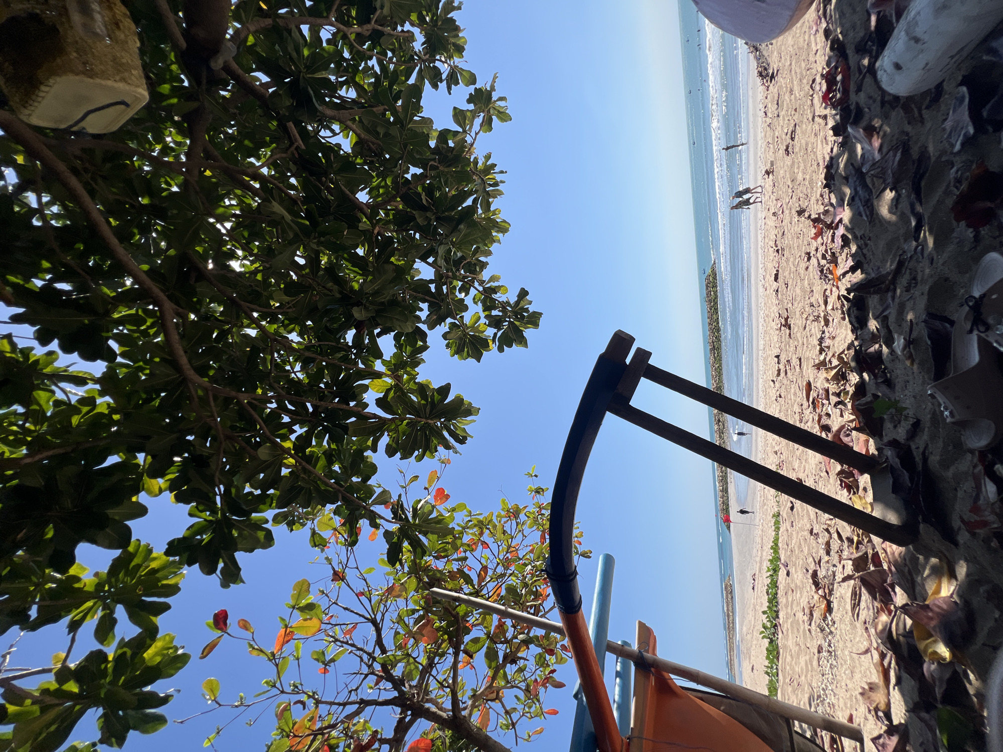 The image depicts a beach scene in Kuta, Indonesia, viewed from under the shade of trees. In the foreground, dried leaves and sand are visible, with parts of a boat or structure and its supports occupying the lower left and right portions of the frame. Beyond this, a sandy beach stretches towards the ocean. Two figures are walking on the beach in the middle ground, with a red flag planted in the sand to their left. In the distance, the ocean meets the horizon under a clear blue sky, with a breakwater visible in the mid-distance. Several small boats are scattered on the water. The overall impression is a sunny day at the beach.