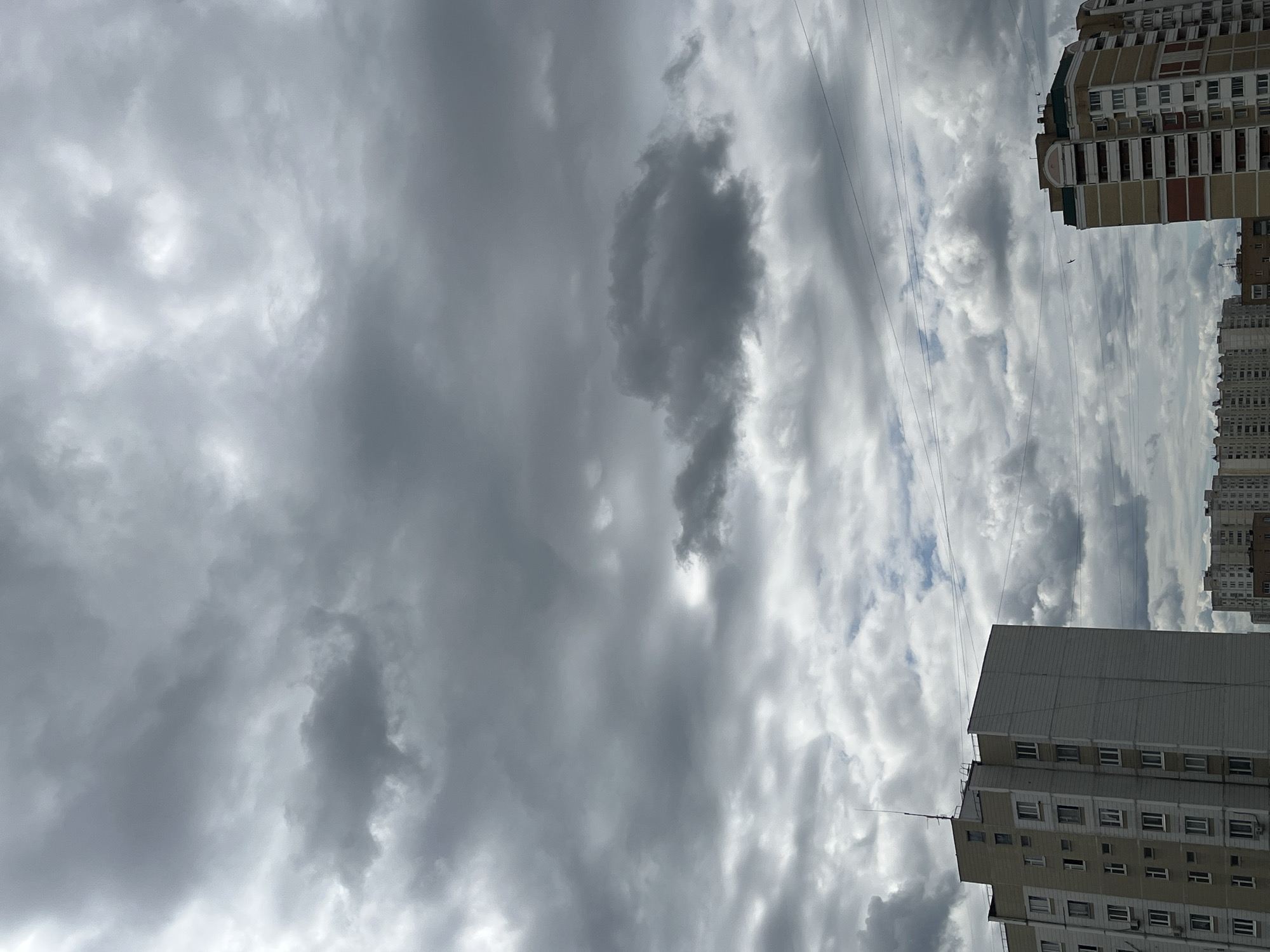 The image depicts a cloudy sky over a city. In the foreground, two apartment buildings are visible. The building on the left is taller and appears to be a standard residential block with multiple stories. The building on the right is also an apartment building with a distinct color pattern on its facade. Several power lines are strung across the sky between the buildings. The sky is filled with dense, dark grey clouds, suggesting an overcast or potentially stormy weather condition. There are no people or specific actions discernible in the image. The scene appears to be a typical urban landscape on a cloudy day.