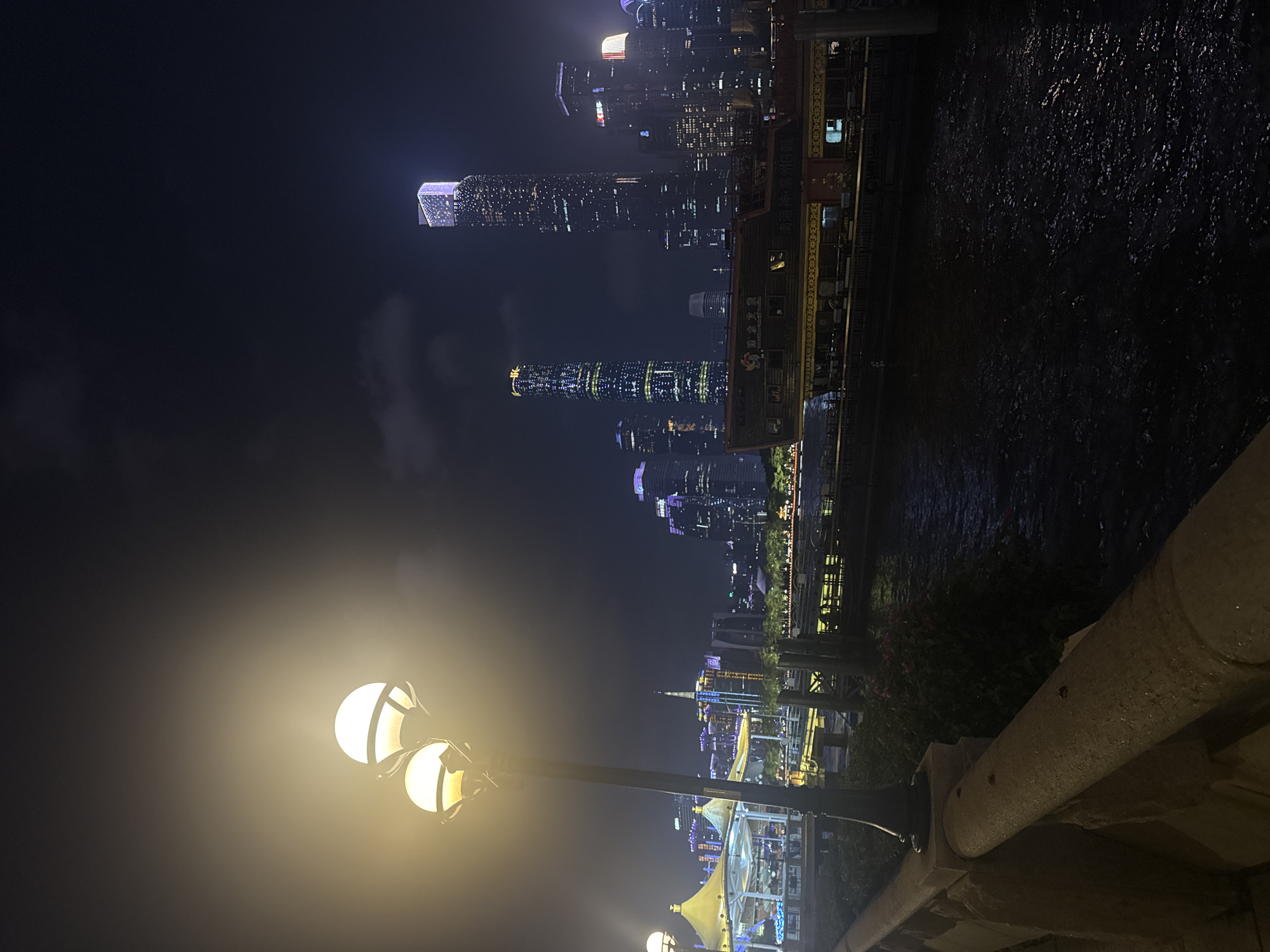 The photograph captures a nighttime cityscape, likely taken from a waterfront promenade in Haizhu District, China. Illuminated modern skyscrapers dominate the background, with distinct architectural styles and varying heights. The Canton Tower is visible. To the right of the skyline is the river, reflecting the city lights.
In the foreground, a multi-bulb street lamp provides a strong source of light. A stone or concrete barrier runs along the bottom right of the frame. The atmosphere is dark and possibly foggy.
