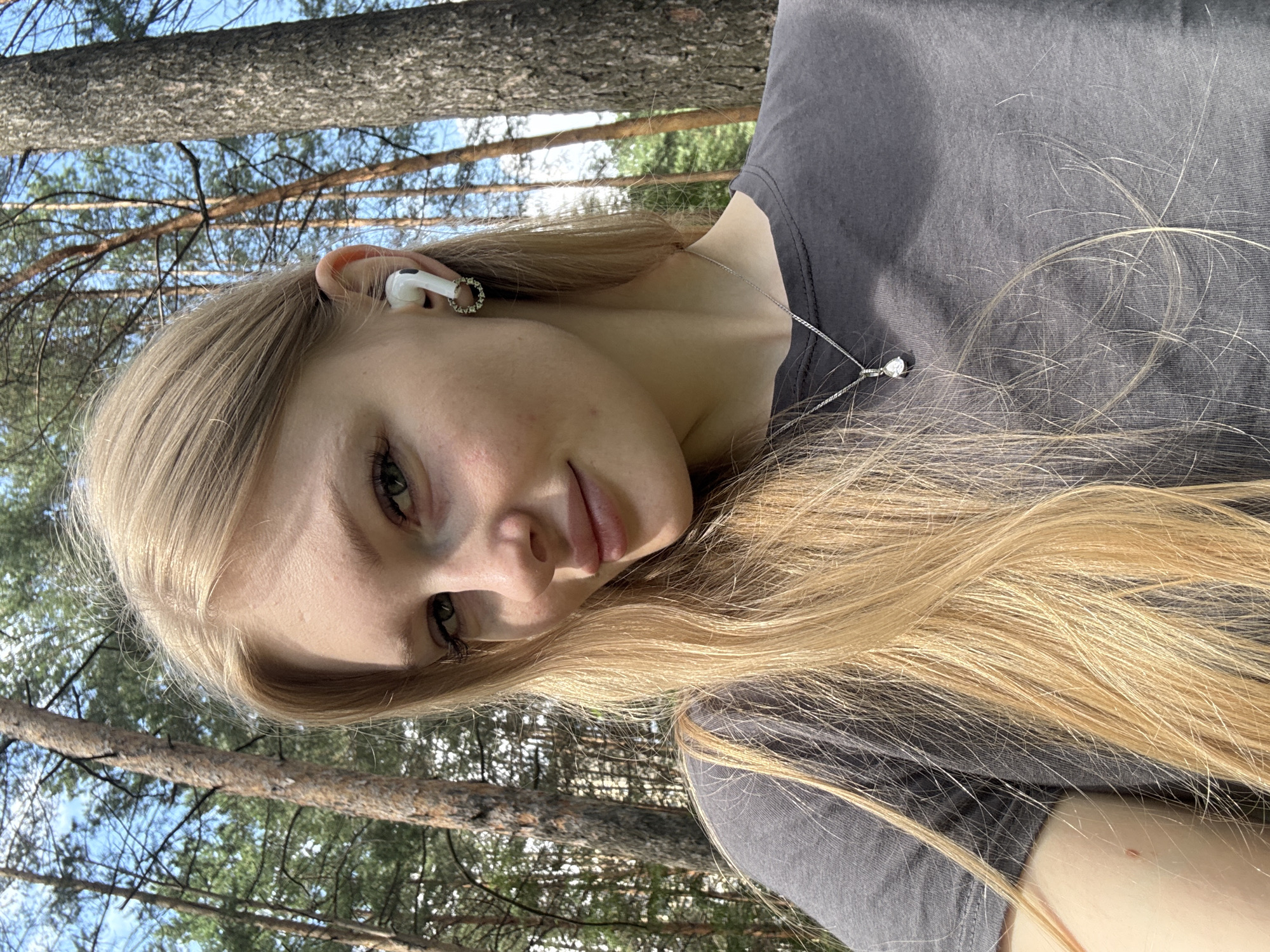 A young woman with blonde hair and green eyes is taking a selfie outdoors, surrounded by trees. She is wearing a dark gray t-shirt and a silver pendant necklace. She has a white earbud in her right ear and a circular earring. The background consists of a forest with tall pine trees and a bright blue sky. The image was taken in the city of Perm, Russia.