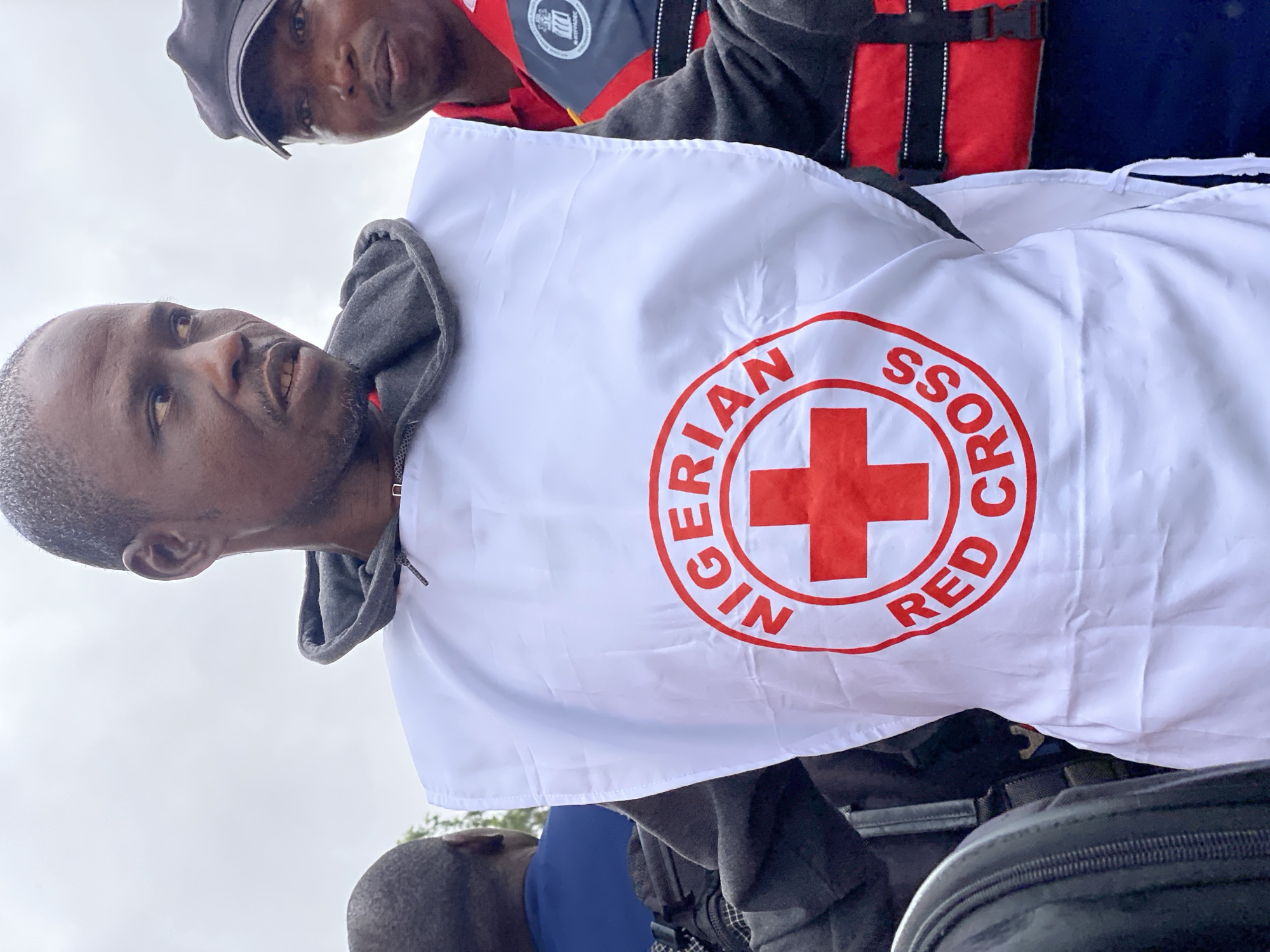 In Kawo, Nigeria, a man wearing a white vest with a red cross and the words "NIGERIAN RED CROSS" is pictured. He has short, dark hair and a mustache. Another man in a black baseball cap stands to his right, and a third person in a blue shirt is partially visible to the left. The sky is overcast.