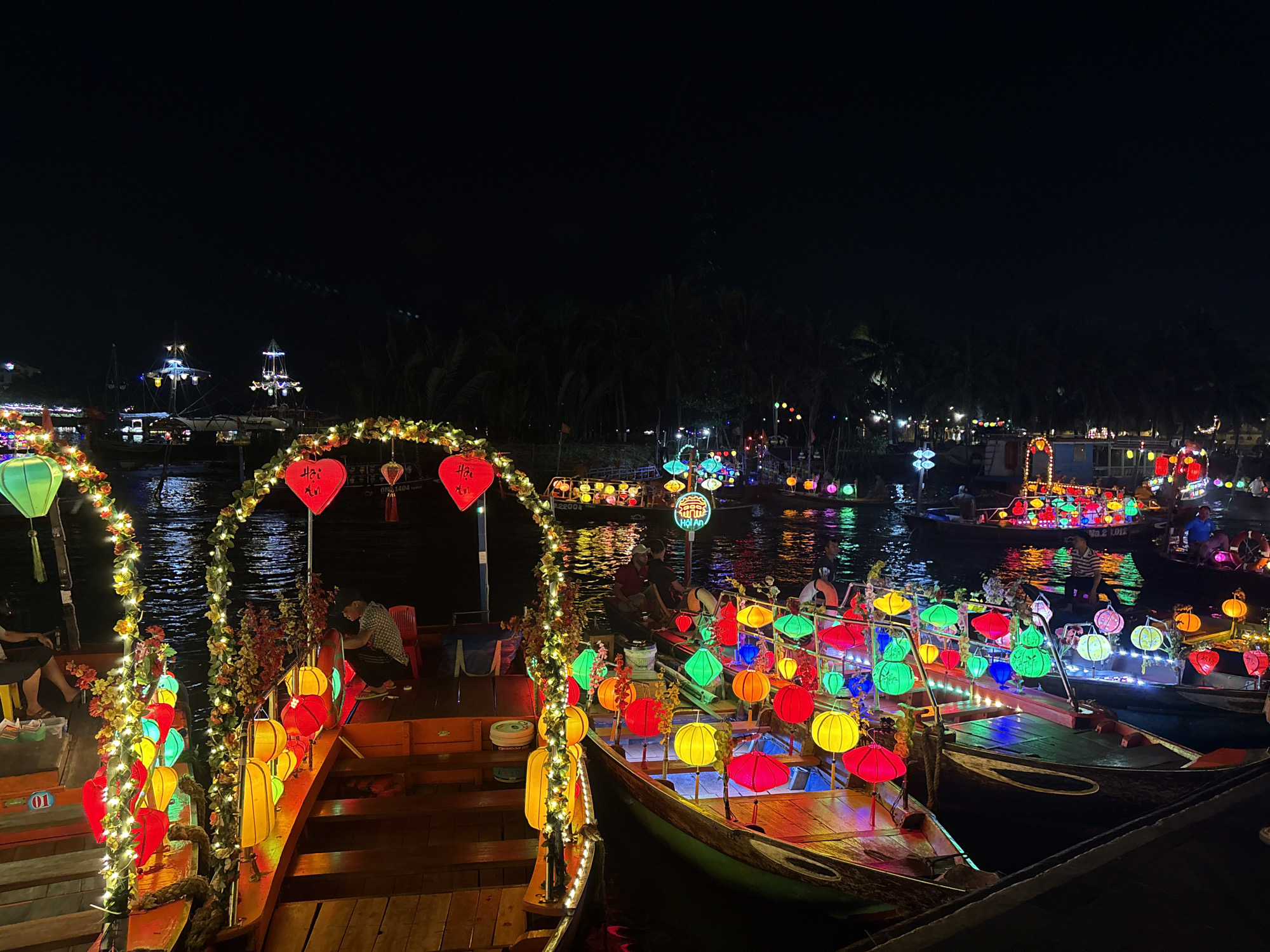 The image shows a night scene on a river in Hội An Ward, Vietnam. Several boats are decorated with colorful lanterns, creating a vibrant display of light. Passengers can be seen on some of the boats. The river reflects the lights, enhancing the visual effect. Lush greenery lines the riverbank in the background.