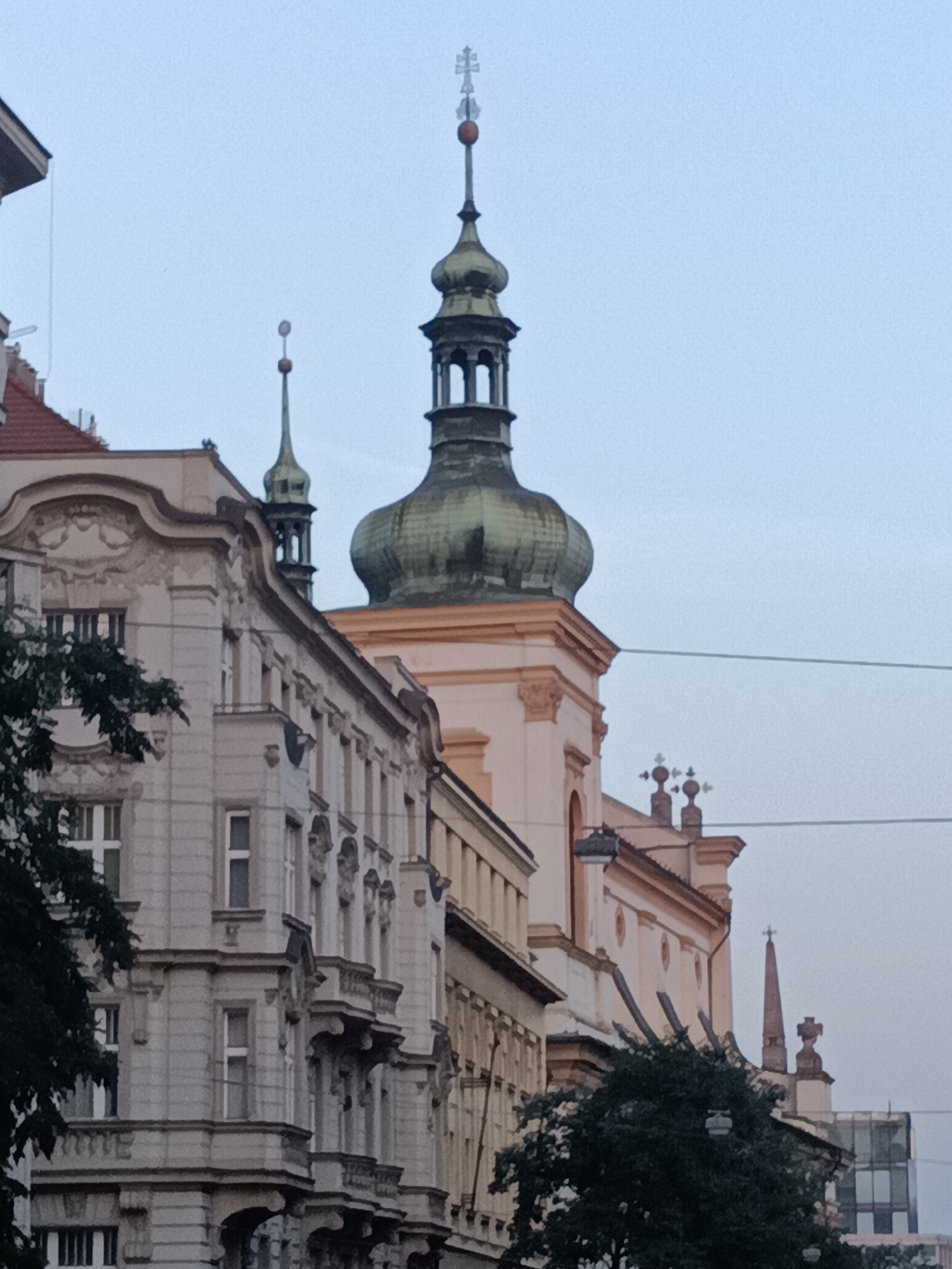 The image shows a view of a street in Prague, Czechia, with several buildings and church spires. The foreground features a multi-story building with ornate architectural details, including balconies and decorative carvings. To the right of this building, the facade of another building with a light-colored exterior and arched windows is visible. Dominating the background are several church spires, one of which has a distinctive green, bulbous dome topped with a cross. The sky is overcast and a light blue-gray color. Electrical wires are visible crisscrossing the upper portion of the frame. Trees with dark green foliage are present in the foreground and mid-ground, partially obscuring some of the buildings. The overall impression is of an urban landscape in a historic European city.