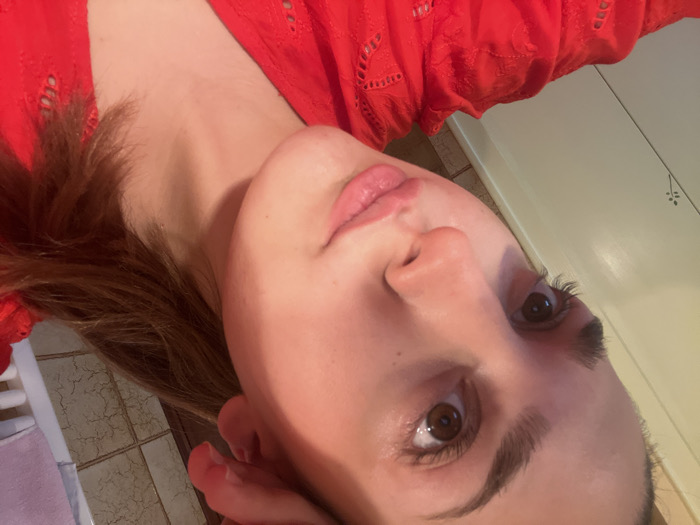 This image depicts a close-up, angled indoor portrait of a female subject, located in Ancenis, France. The subject, a young adult with fair complexion, brown eyes, and medium-brown hair, is oriented facing slightly upward and to the viewer's left. Her lips are lightly parted. She is wearing a red garment featuring a patterned, possibly eyelet, design. Her right hand is positioned near her right ear/temple area, partially obscured. The background elements include a light-colored, possibly cream-colored, vertical panel on the left, and light brown square-tiled walls on the right. A white, possibly tubular, fixture, consistent with a radiator or towel warmer, is partially visible in the upper right background. The lighting appears artificial and warm.