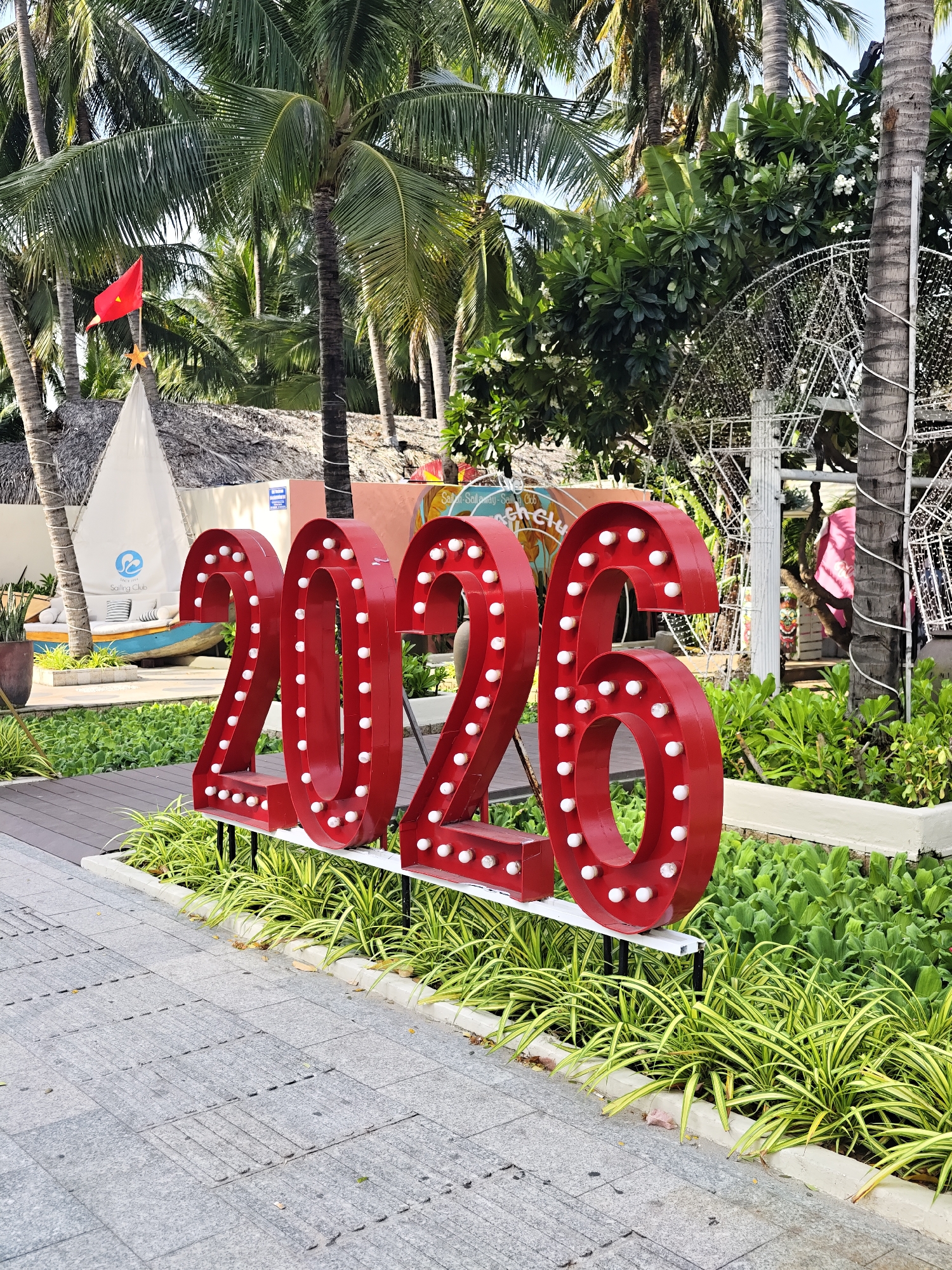 The image shows a large red sign with white lights spelling out "2026". The sign is in front of lush green foliage and palm trees. Behind the sign, there is a white structure resembling a sailboat with a thatched roof. A red flag is visible on top of the sailboat structure. The ground in front of the sign is a paved walkway. The scene appears to be outdoors, likely in a tropical location. The image is focused on the "2026" sign. The overall impression is festive and possibly related to a celebration or event.