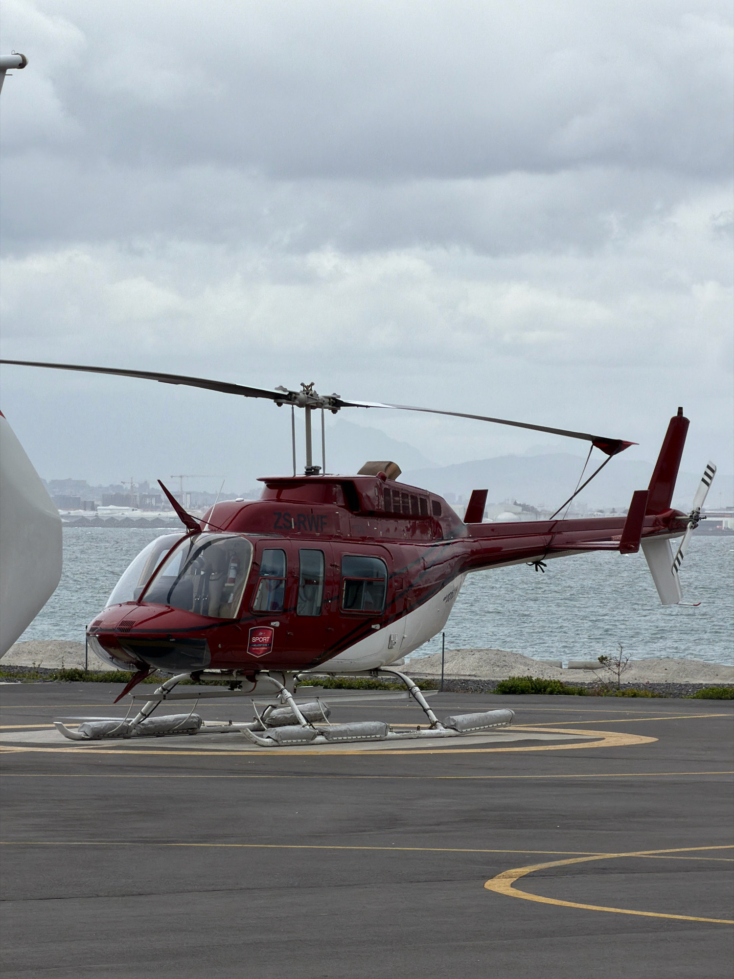 An outdoor, full shot captures a dark red and white helicopter situated on a helipad. The helicopter is oriented facing the viewer's right, with its rotor blades extended horizontally. The helipad is marked with yellow lines and is paved with asphalt. In the background, the ocean meets a shoreline with some vegetation and distant hills. The sky is overcast and appears grey. The helicopter bears the registration number ZS-RWF and features a "SPORT HELICOPTERS" logo on its side. Another helicopter, mostly white, is partially visible on the left side of the frame. The scene is static, with no people or apparent actions taking place. The overall impression is one of a functional helipad and aircraft under a cloudy sky. The location is the V&A Waterfront in Cape Town, South Africa, which is a popular tourist destination with a marina, hotels, and recreational activities, as well as a heliport.