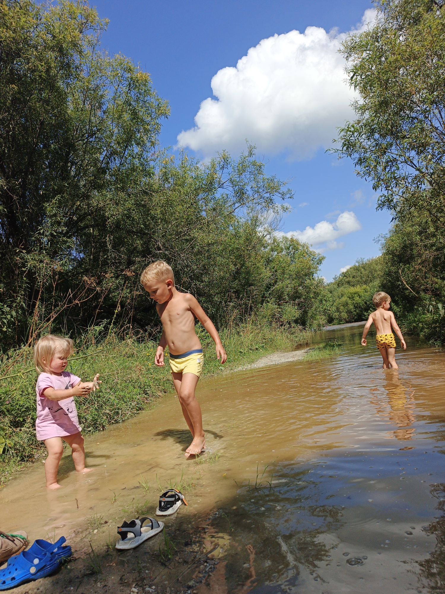 Two young children are playing in a shallow, muddy body of water. A blonde-haired boy, wearing yellow shorts, stands facing the viewer and looking down. Beside him, a younger blonde-haired girl in a pink t-shirt is holding up her hands, which appear to be covered in mud. To the right and further back in the water, another blonde-haired boy in yellow shorts walks away from the camera. The environment is natural, with lush green trees and vegetation lining the banks of the water. The sky is blue with fluffy white clouds. Several pairs of sandals and a blue clog are visible on the muddy bank in the foreground. The scene appears to be a natural outdoor location, potentially a river or stream, in or near the village of Medvedyovka, Russia.