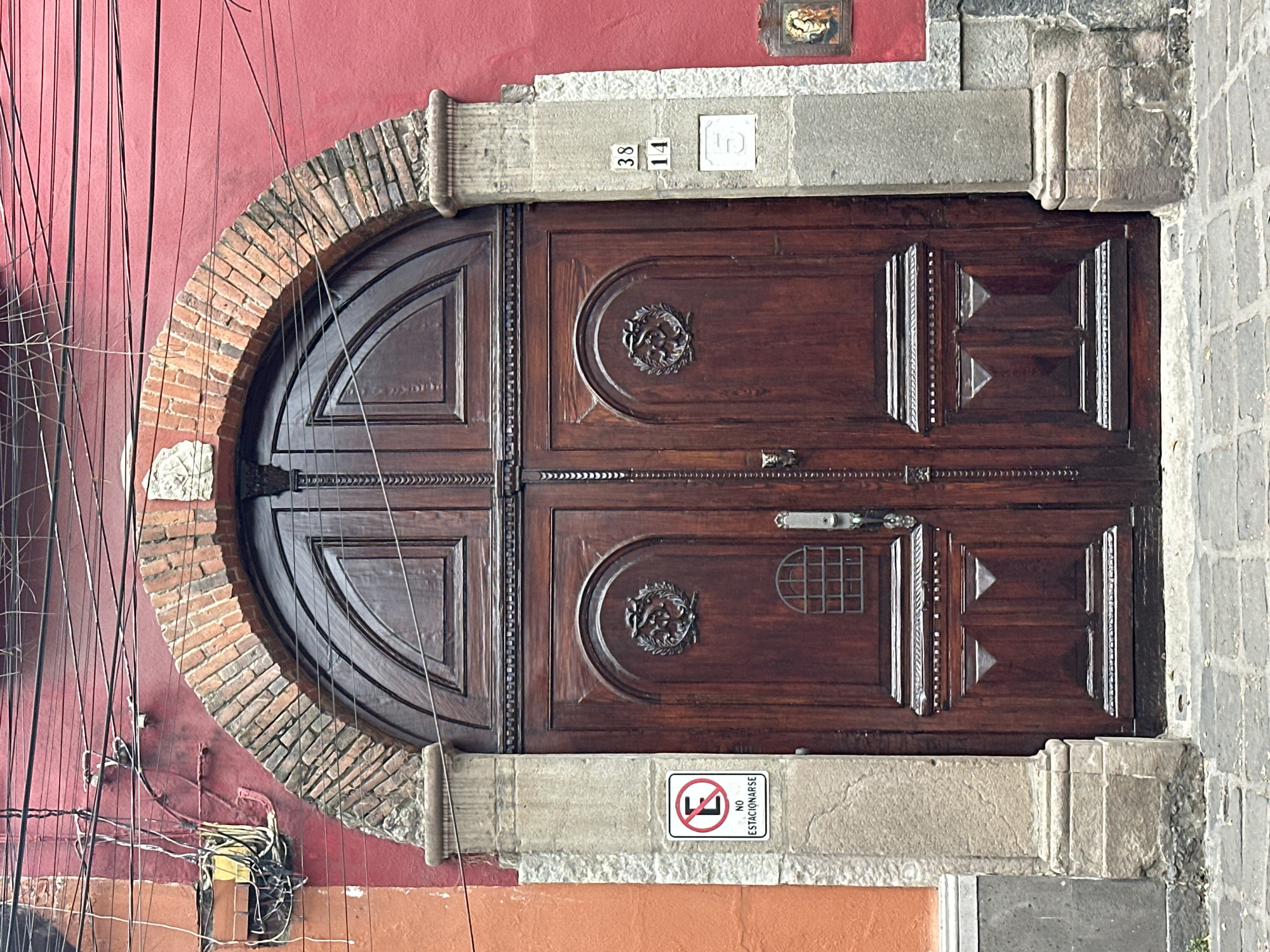 A close-up, slightly angled shot captures a large, ornate wooden door with an arched brick frame set into a brightly painted reddish-orange wall. The door is composed of two panels, each decorated with raised geometric designs and circular insets featuring ornate metal medallions. A metal handle and what appears to be a small viewing grille are visible on the right panel. Above the door, the stone lintel bears the numbers "38" and "14", and another unreadable sign, possibly indicating the address. Below the door on the left side, a parking restriction sign in Spanish reads "NO ESTACIONARSE" (No Parking). The facade of the building is visible on both sides of the door, showing color variations in the paint. A dense network of electrical or communication wires hangs to the left of the door. The location is assumed to be Mexico City, Mexico.