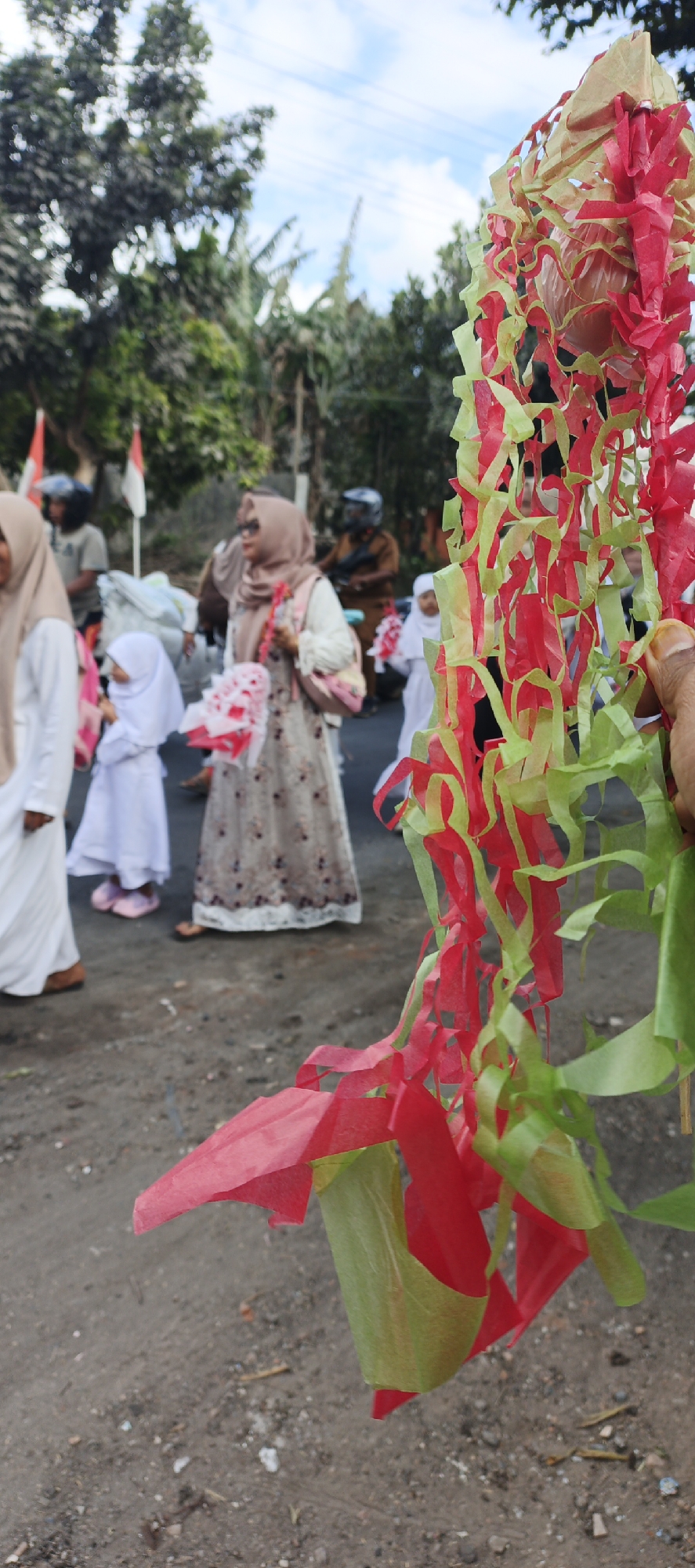 In Grenden, Indonesia, a blurry outdoor scene unfolds with a focus on festive decorations and a procession of people. In the foreground, a person's hand holds a cluster of red and green crepe paper streamers, possibly part of a celebratory garland or decoration, obscuring the view of a paved road and dirt ground. In the mid-ground, several individuals are walking, including two young girls in white hijabs and dresses, each holding a small bouquet of pink and white flowers. A woman in a patterned dress and hijab walks between them, also carrying a bouquet. Behind them, other people and a motorcycle with a rider wearing a helmet are visible, as well as Indonesian flags. The background features trees and a cloudy sky, suggesting an overcast day.
