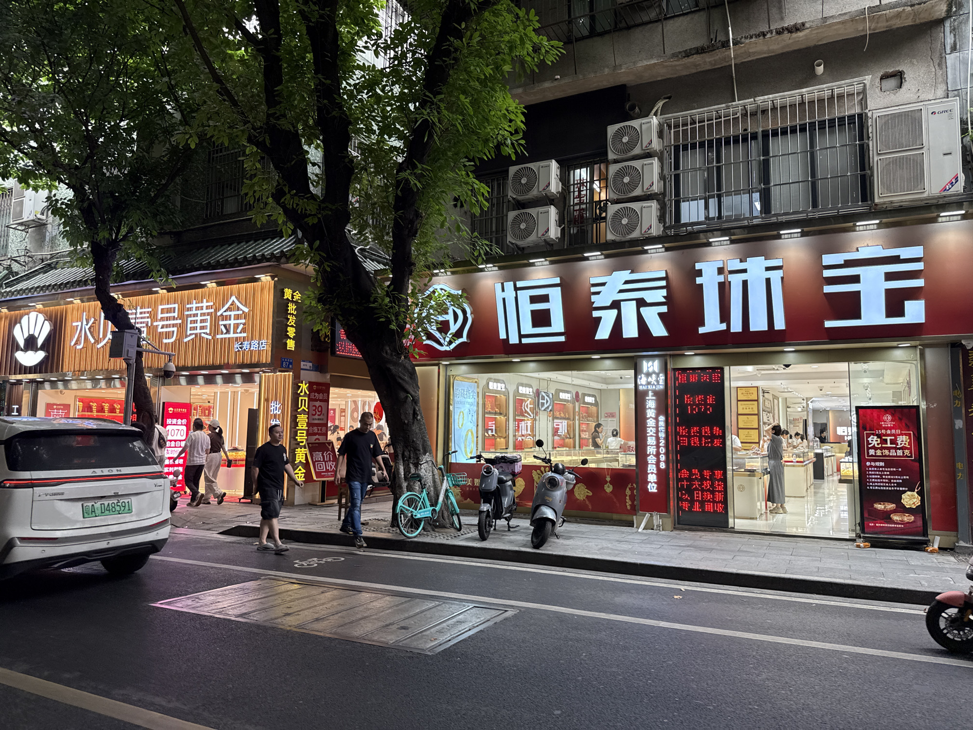 The image depicts a street scene in the Liwan District of Guangzhou, China. On the right, a jewelry store named "Hengtai Jewellery" with illuminated Chinese characters stands next to another gold retailer, "Shui Bei Yi Hao Gold." The storefronts are adorned with promotional signs and feature bright lighting.
In front of the stores, a man in a black shirt and jeans walks along the sidewalk, and another man appears to be locking a blue bicycle to a tree. Two electric scooters are parked next to the bicycle.
A white AION electric vehicle is parked along the curb, bearing the license plate "A D48591." Other people are visible walking in and out of the stores.
The buildings are multi-story, and several air conditioning units are attached to the exterior of the upper floors. A tree with dense foliage casts shadows across the scene. The street is partially visible, and street lighting provides illumination.