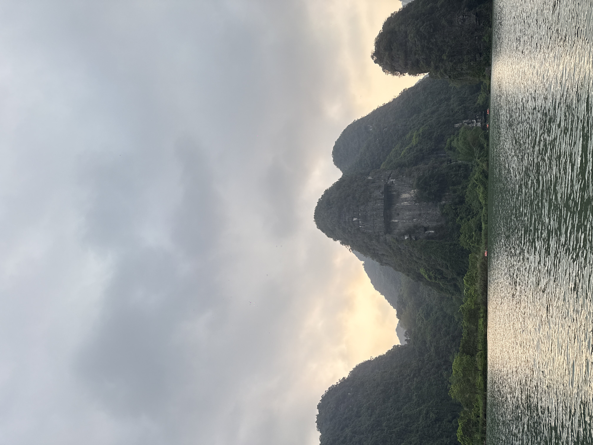 The image depicts a scenic view in Hoa Lư Ward, Vietnam. On the right side is a body of water with visible ripples, reflecting sunlight. A line of greenery, possibly trees or vegetation, separates the water from the land. In the background are karst mountains, their peaks covered in lush green vegetation. The sky is overcast with grey and white clouds, and a soft, golden glow is visible behind the mountains, suggesting either sunrise or sunset. Several small boats are visible on the water near the base of one of the mountains. The overall impression is one of natural beauty in a humid, tropical environment.