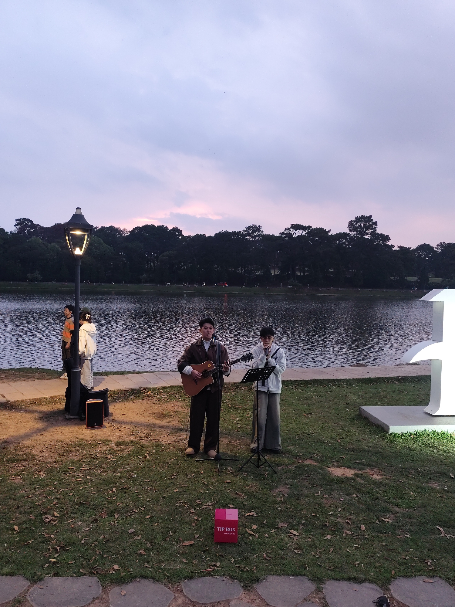 The image captures an outdoor musical performance by a lake in Da Lat, Vietnam, during twilight. In the foreground, two young men are performing on a grassy patch. One, positioned to the left, plays an acoustic guitar and sings into a microphone, dressed in a dark jacket. To his right, the other man sings into a microphone on a stand, wearing a light-colored hoodie and wide-leg pants. A small black speaker or amplifier rests on the ground near the guitarist.
A red "TIP BOX THANK YOU" is prominently displayed on the grass in the lower-left corner, indicating a busking or street performance. Behind the performers, a paved walkway runs along the edge of a calm lake, whose surface reflects the fading light. The opposite bank is lined with a dense forest of trees, extending across the horizon.
On the left, a lit lamppost illuminates the area, near which two or three onlookers stand. To the right, a large, illuminated white sculptural element, possibly a stylized '1' or part of a larger artwork, adds to the evening ambiance. The sky above transitions from a soft blue to purplish and pink hues, characteristic of dusk. The scene conveys a peaceful and artistic evening by the lake.