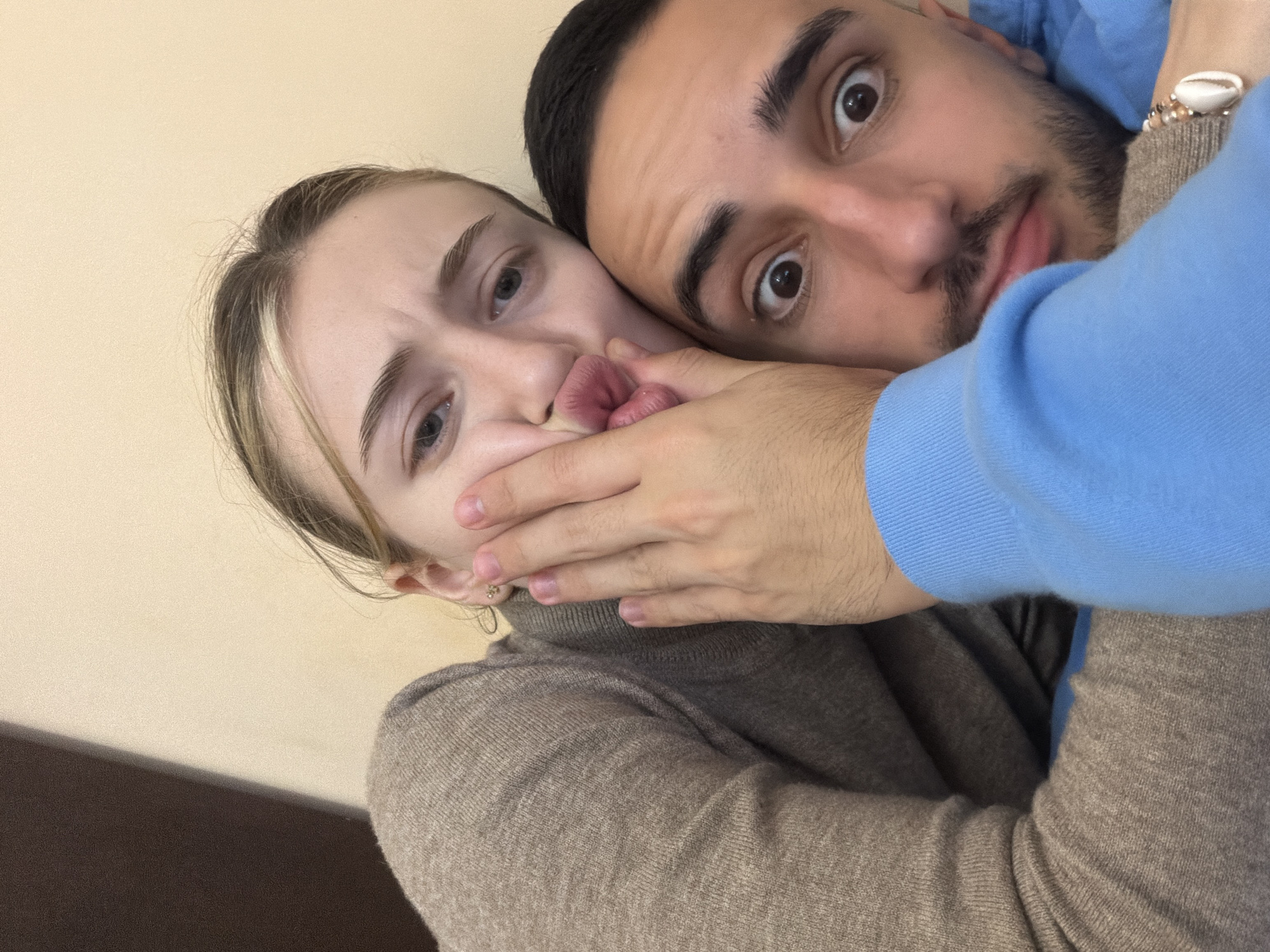 A close-up, angled photograph captures two individuals: a young woman and a young man. The woman, positioned on the left, is wearing a light brown turtleneck sweater. Her mouth is covered by the man's hand, and she appears to be grimacing. The man is leaning in from the right, with his face close to the camera. He has dark hair, a beard, and wide eyes. He is wearing a blue long-sleeved shirt over a brown sweater. The background is a plain, light beige wall. A dark-colored wooden surface is visible in the lower-left corner. No elements indicate the location of Plovdiv, Bulgaria.