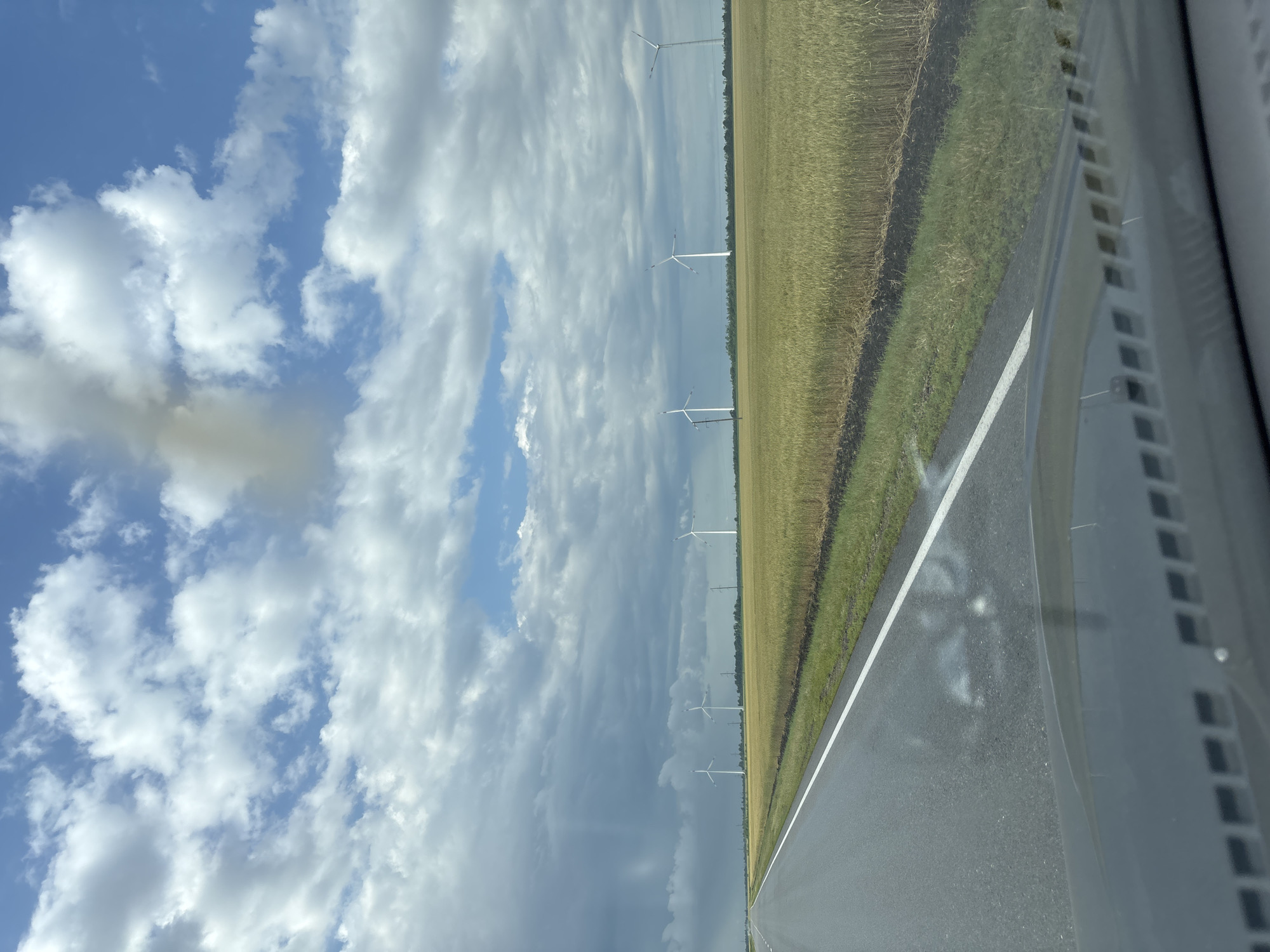 The image depicts a wide, asphalt road stretching into the distance. A white line marks the right edge of the road. To the right of the road, there is a field of dry, golden grass, likely a harvested crop. Beyond the field, a line of white wind turbines stands against a dramatic sky. The sky is a mixture of bright blue with fluffy white clouds and darker, overcast areas with patches of blue peeking through. The clouds suggest a dynamic weather system, possibly with impending rain or recent showers. The scene appears to be set in a rural, open landscape. The perspective is from inside a vehicle, with the dashboard and windshield visible in the lower right corner. The light suggests it is daytime, likely late morning or afternoon. There is no visible text in the image. The overall mood is one of openness and the vastness of nature, with the human-made structures of the road and wind turbines interspersed within the landscape.
