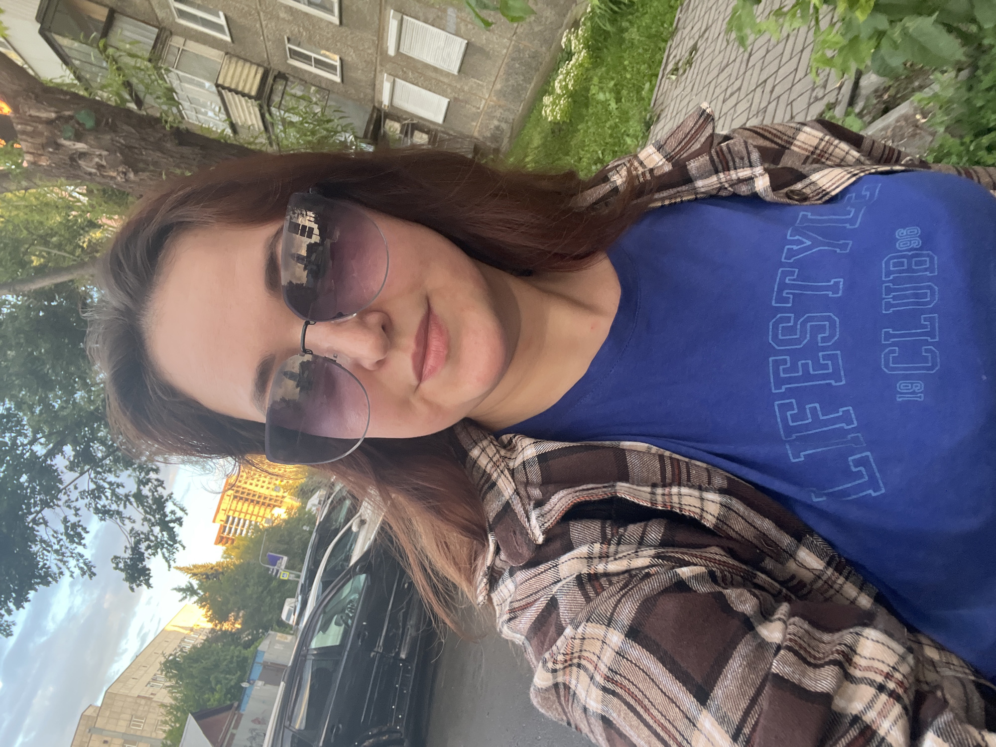 A medium-shot, selfie-style photo captures a woman with medium-length brown hair and sunglasses, wearing a brown and tan plaid shirt over a blue t-shirt with "LIFESTYLE CLUB 96" printed on it. She is standing outdoors in an urban environment, with a sidewalk and green grass to her right. In the background, parked cars, a tree, and buildings are visible. The image appears to have been taken in Chelyabinsk, Russia, based on the architectural style of the buildings and the overall urban setting, consistent with residential areas found in the city. The lighting suggests it is late afternoon or early evening, with the sun casting a warm glow on the buildings.