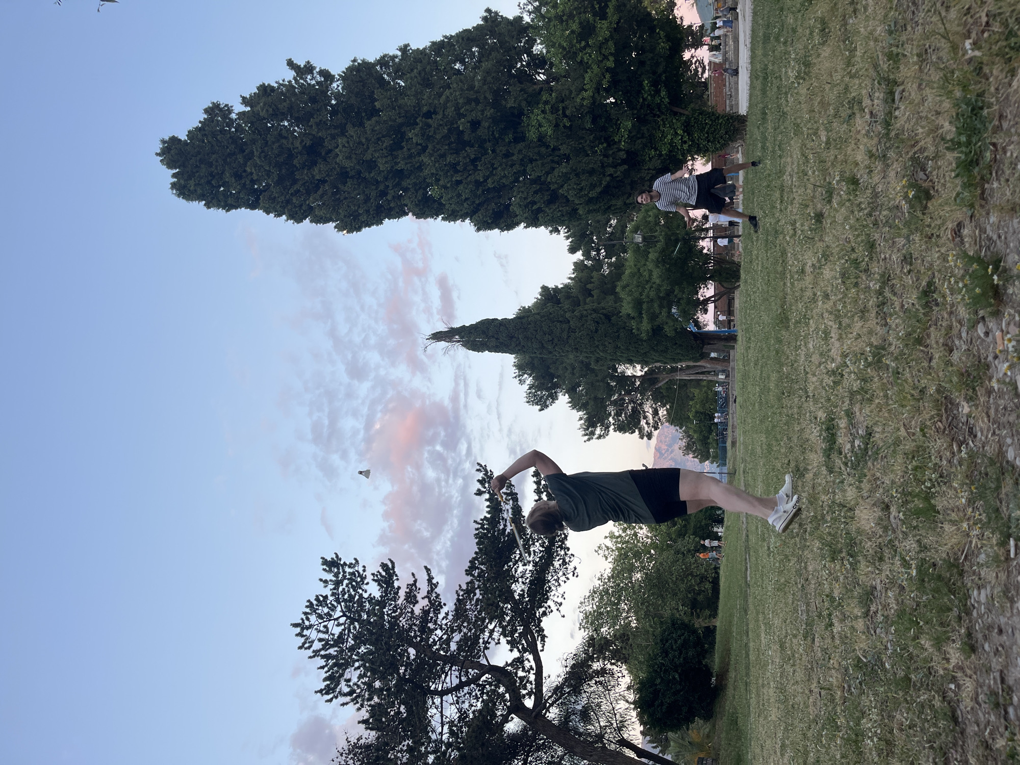 The photograph shows two individuals playing badminton on a grassy area in Budva, Montenegro. In the foreground, a person wearing a green t-shirt and black shorts is mid-swing with a badminton racket, presumably hitting a shuttlecock in the air. A second individual in a striped shirt and dark shorts is standing further back, near a line of trees. The sky is a mix of blue and pink, suggesting it is either dawn or dusk. The grassy area has a mix of green and dried grass. Trees of varying sizes and types are scattered in the background.