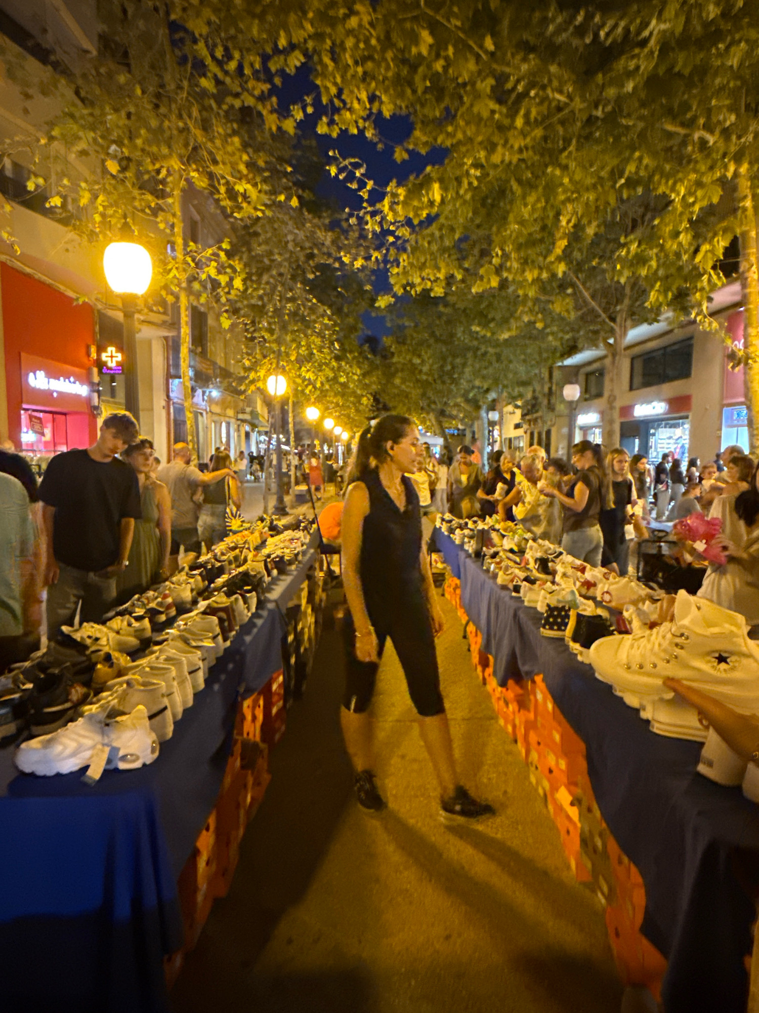 The image depicts a street market at night in Blanes, Spain, likely on a commercial street with trees lining the sidewalk and lampposts illuminating the scene. The market features numerous tables covered in blue tablecloths, displaying a large variety of footwear, primarily sneakers. The footwear is neatly arranged in rows, with some pairs on display and others in boxes underneath.
Several individuals are present, browsing the merchandise and interacting with each other. In the foreground, a woman in dark athletic wear is walking past a table of shoes. To the left, a young man in a black t-shirt and jeans stands with a woman in a green dress, both looking towards the market. Other people are scattered throughout the scene, suggesting a bustling atmosphere. The background shows buildings with illuminated storefronts, indicating the market is set up along a street in a town or city. The overall impression is of a lively evening market scene.
