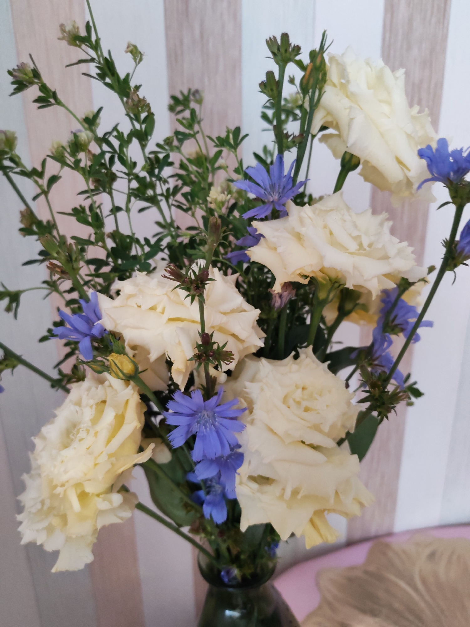 A vertical, medium shot shows a bouquet of creamy white roses and blue chicory flowers in a dark green glass vase. The bouquet is centrally placed and takes up most of the frame. The background is a wall with vertical, pale wood-effect panels alternating with white stripes. To the right of the frame, a pink surface is visible, with what appears to be a light brown, fuzzy object resting on it. The lighting is diffused and natural, coming from the front. The scene appears to be indoors, possibly in a domestic setting in Izhevsk, Russia. There are no people present in the image.