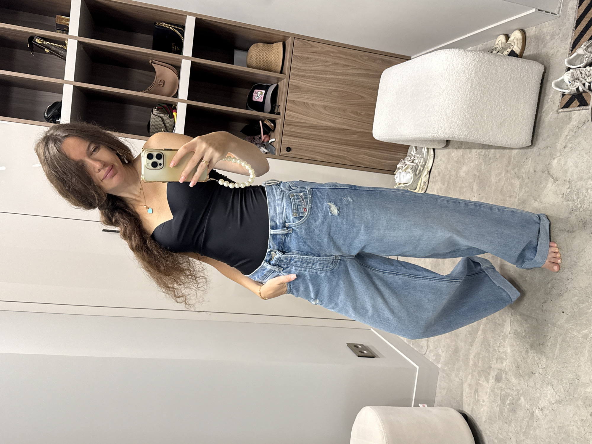 A woman with long brown wavy hair is taking a selfie in a room with a minimalist interior. She is wearing a black strapless top and light-wash, high-waisted, wide-leg jeans that are rolled up at the ankles. She has a pearl necklace and a turquoise pendant necklace on. Her left hand is in her pocket, and she is holding a gold smartphone in her right hand. Behind her is a tall wooden shelving unit filled with various handbags and hats. To her left, there is a small round white ottoman. To her right, there is a larger white textured ottoman. Several pairs of sneakers are visible on the floor around the ottomans. The overall scene suggests a personal dressing area or a fitting room in a boutique. The location in the city of Dolgoprudny, Russia, is not discernible from the visual information provided in the image.