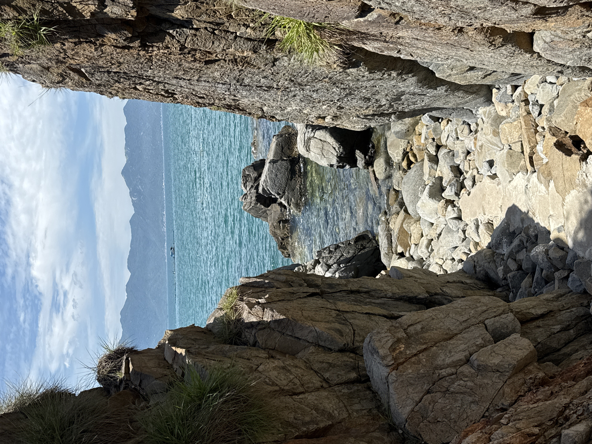 This is a vertical, medium shot photograph taken on a sunny day, depicting a rocky coastal path leading to the turquoise waters of the sea in Vĩnh Lương, Vietnam. The path is narrow and bordered by large, rough, gray and brown rock formations. Some sparse green grass grows out of crevices in the rocks. The sea in the distance is calm with small waves. In the background, a range of hazy blue mountains meets the horizon under a sky with scattered white clouds. A small, distant boat with a white roof is visible on the water. The foreground and midground are dominated by the textured rocks and the rocky shore.