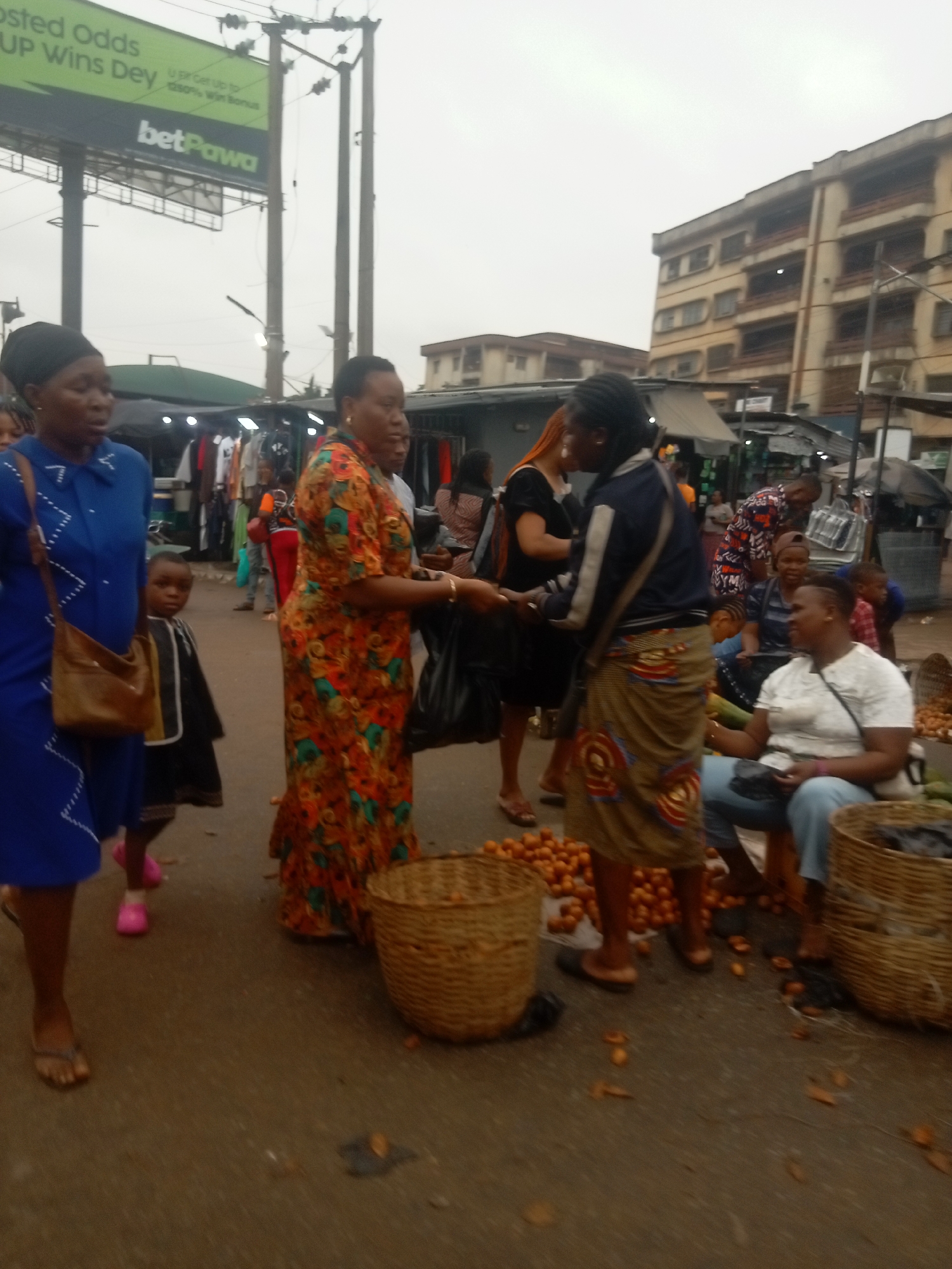 The image depicts a bustling street scene in Onitsha, Nigeria. Several people are present, engaged in commerce. A woman in a blue dress and a brown purse walks to the left, while another woman in an orange floral dress appears to be handing something to a third woman wearing a jacket and a patterned wrap. These interactions occur near baskets full of oranges. In the background, buildings and market stalls are visible, with a large "betPawa" billboard overhead. The weather appears overcast, casting a muted light on the scene.