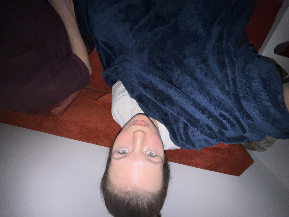 A person lies upside down on an orange couch, partially covered by a navy blue blanket. They wear a white shirt. A burgundy-colored object is on the left side of the couch. The person has fair skin, green eyes, and brown hair pulled back. The background consists of a white wall and a glimpse of another object in the top right corner.