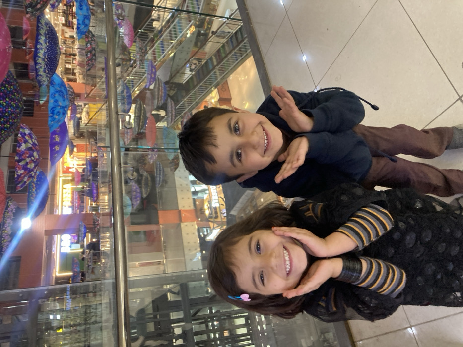 The image captures two smiling children, a girl and a boy, posing in an indoor multi-story shopping mall located in Quetta City Tehsil, Pakistan. The girl on the left, with dark hair and a pink hair clip, wears a black textured long-sleeved top over striped undersleeves and multiple black bangles on her wrist, with her hands positioned playfully on her cheeks. The boy on the right, also with dark hair, is dressed in a dark blue hooded sweatshirt and brown trousers, holding his hands open and raised near his chest. Both children are looking directly at the camera with broad smiles. Behind them, a glass safety barrier overlooks the lower levels of the mall. The background reveals a modern commercial environment with multiple escalators, various illuminated shop signs including "MAZZA" and "SVERTO," and a prominent overhead decoration consisting of numerous inverted, colorful, patterned umbrellas suspended from the ceiling. The floor is light-colored tile, and architectural elements include glass panels, reflective surfaces, and vertical wall sections. The scene is brightly lit by artificial light sources.