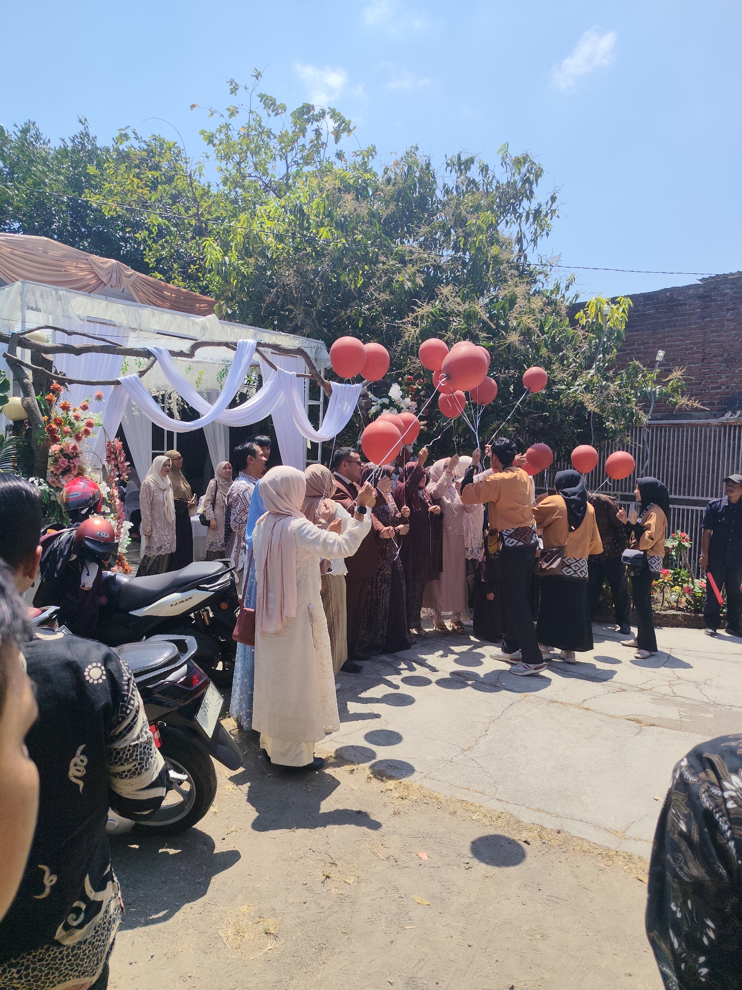 This image captures an outdoor scene in what appears to be a residential area in Garut, Indonesia, possibly during a celebration. A group of people, mostly women dressed in traditional attire including hijabs, are gathered under a makeshift canopy decorated with white drapes and pink flowers. Several individuals are holding red balloons, with some appearing to be releasing them. In the foreground, a motorcycle is parked, and parts of other people are visible at the edges of the frame. The environment includes a brick wall on the right and lush green trees and a bright blue sky above, suggesting a sunny day. The ground is paved with concrete in some areas and dirt in others.