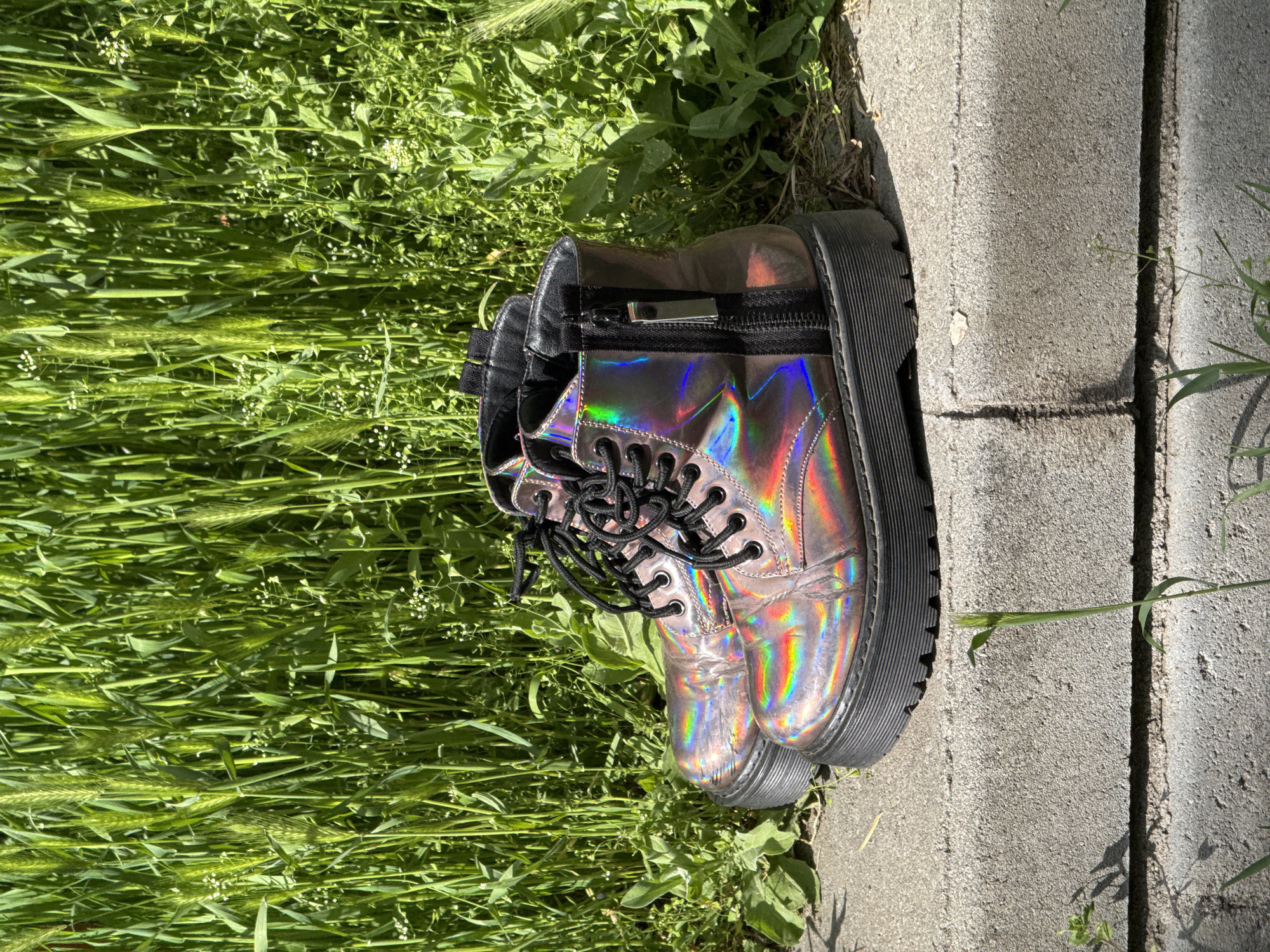 The image shows a pair of iridescent, laced boots placed on the edge of a concrete walkway. One side of the boots borders tall, green grass. The boots are black-laced and have a thick, black sole. The walkway is made of textured concrete slabs with visible seams. The scene is outdoors and is brightly lit. There is no indication that the photo was taken in Sofia, Bulgaria. The specific location is not identifiable from the image.