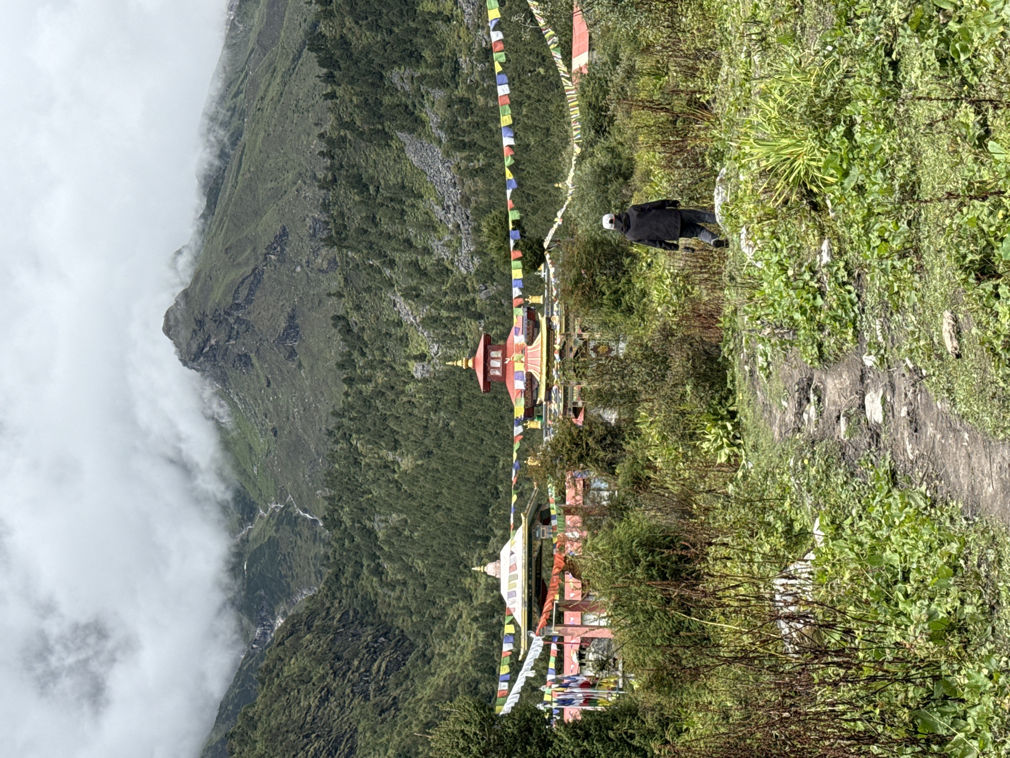 A person wearing a dark jacket and a white cap walks away from the camera on a dirt path through lush green vegetation. The path leads towards a colorful Buddhist monastery complex, characterized by vibrant prayer flags strung across the scene. Behind the monastery, steep, forested mountains rise, partially obscured by low-lying clouds, with a waterfall visible cascading down the mountainside. The overall environment is mountainous and appears to be in a remote, natural setting, likely Samagaon, Nepal.