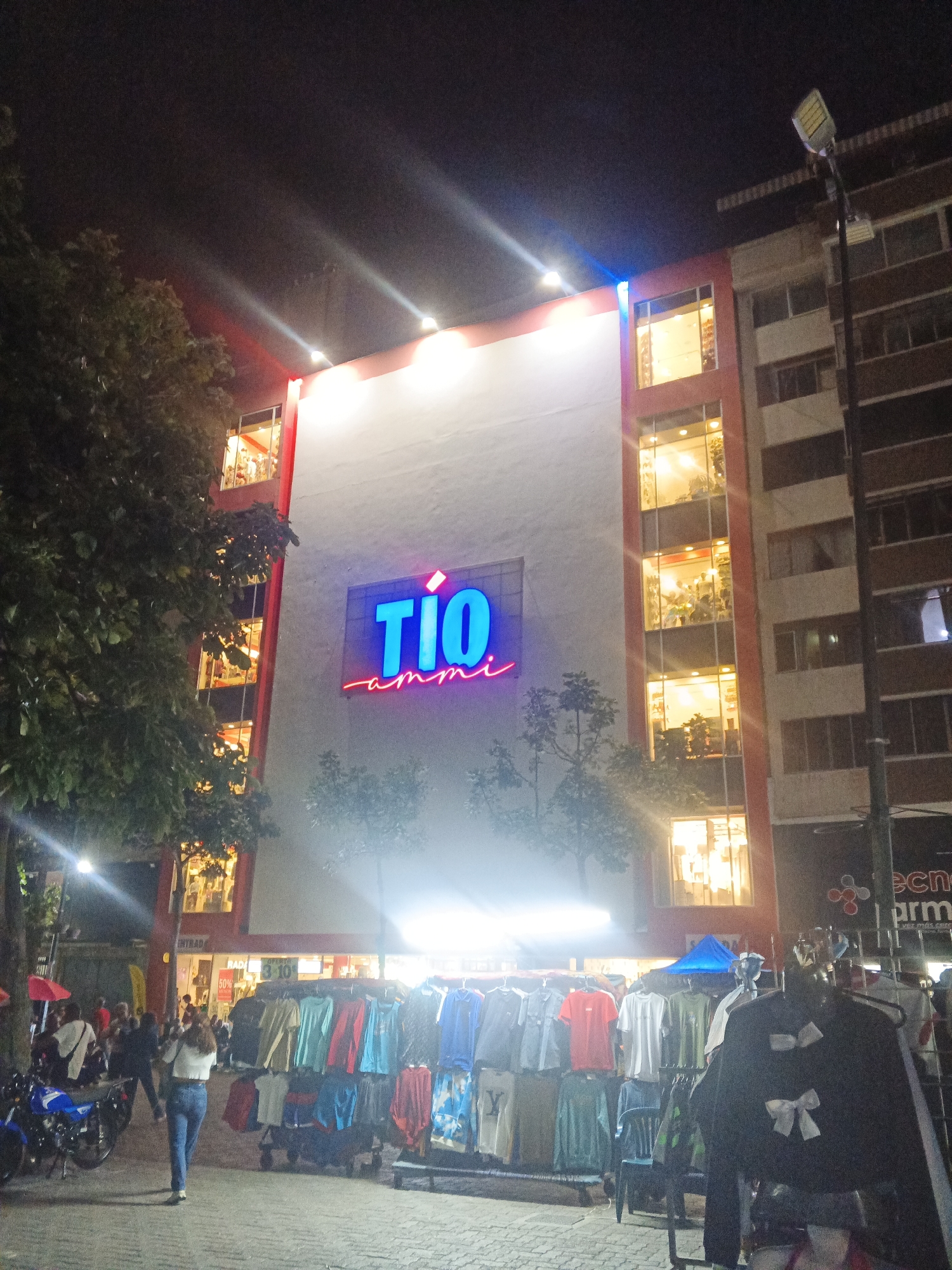 The image shows a busy street scene at night in Caracas, Venezuela. A large building with a prominent "TIO AMMI" sign is lit up, showcasing multiple floors of interior space. The street in front of the building is lined with stalls selling various clothing items, and people are browsing the merchandise. A woman in a white top and blue jeans walks away from the camera, while a blue motorcycle is parked on the left. Trees and streetlights are also visible, contributing to the urban night atmosphere.