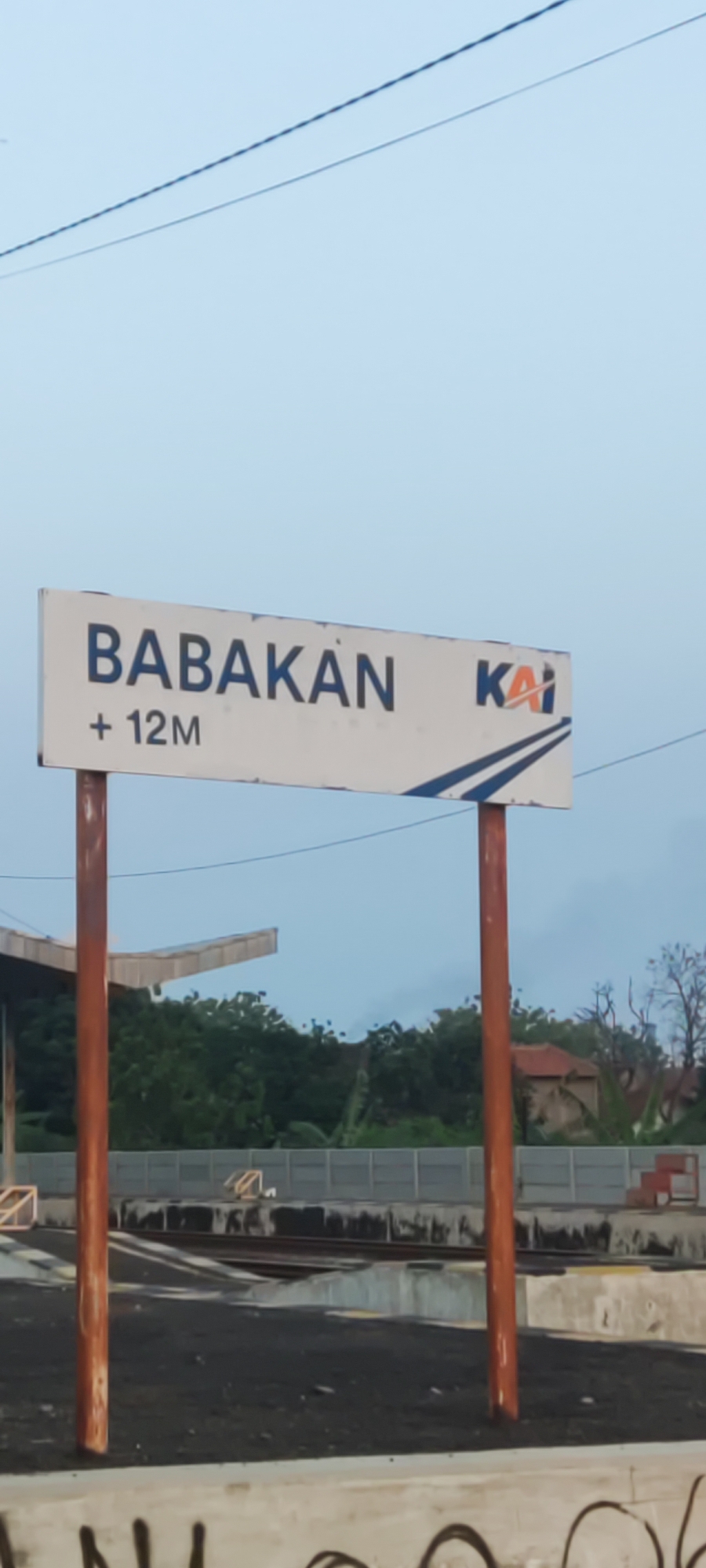 The image shows a sign for the "BABAKAN + 12M" train station, identified by the "KAI" logo, which stands for Kereta Api Indonesia (Indonesian Railways). The sign is white with dark blue lettering and a blue graphic of train tracks. It is mounted on two rusty metal poles. In the background, there are train tracks, a platform, trees, and some buildings. The sky is overcast. The location is in Pakusamben, Indonesia.