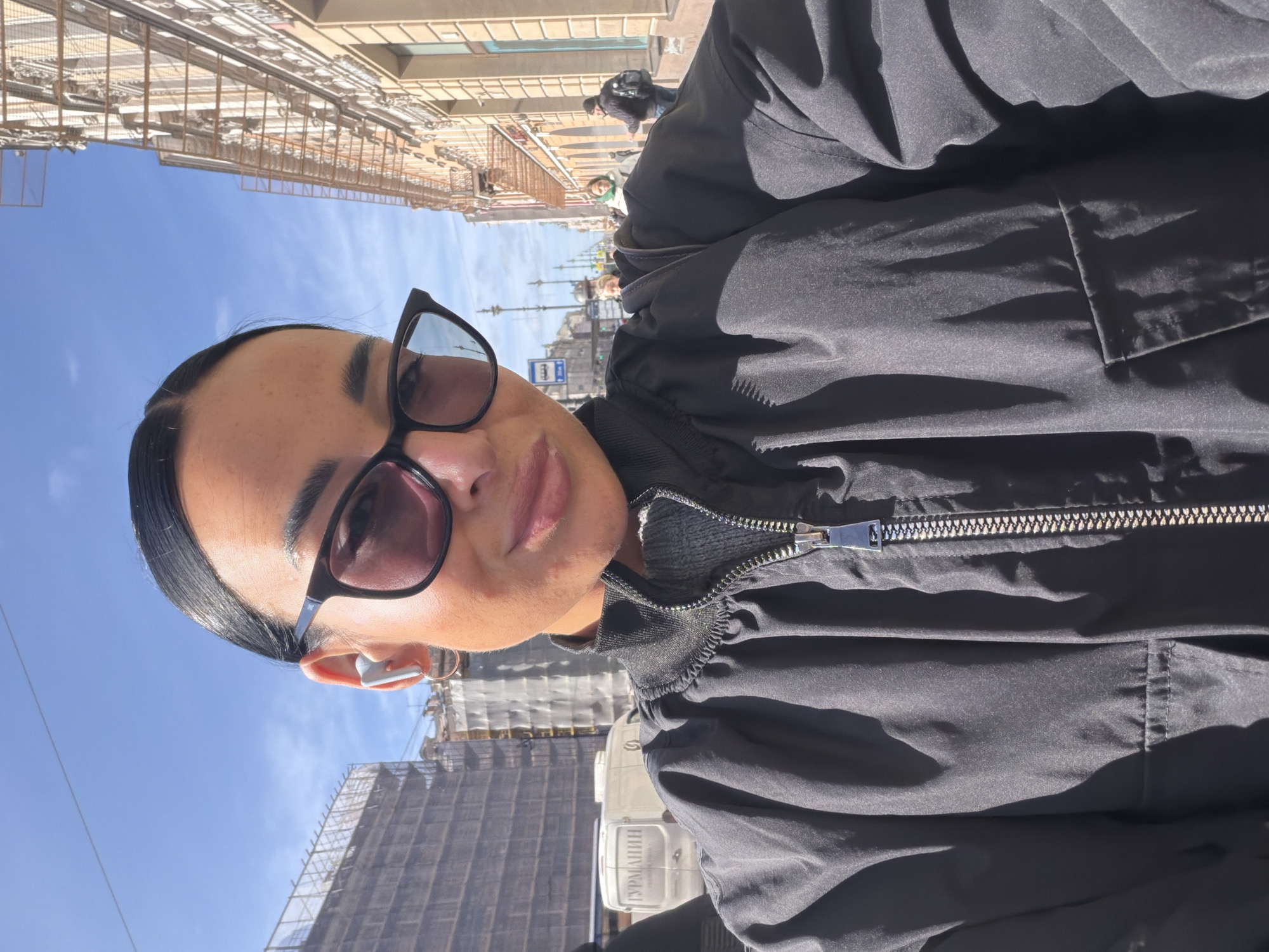 The image shows a person wearing sunglasses and a black jacket, standing on a street in Saint Petersburg, Russia. Buildings with scaffolding are visible in the background, as well as a blue sky. The person is wearing an earpiece in their left ear. Other people are walking in the distance.
