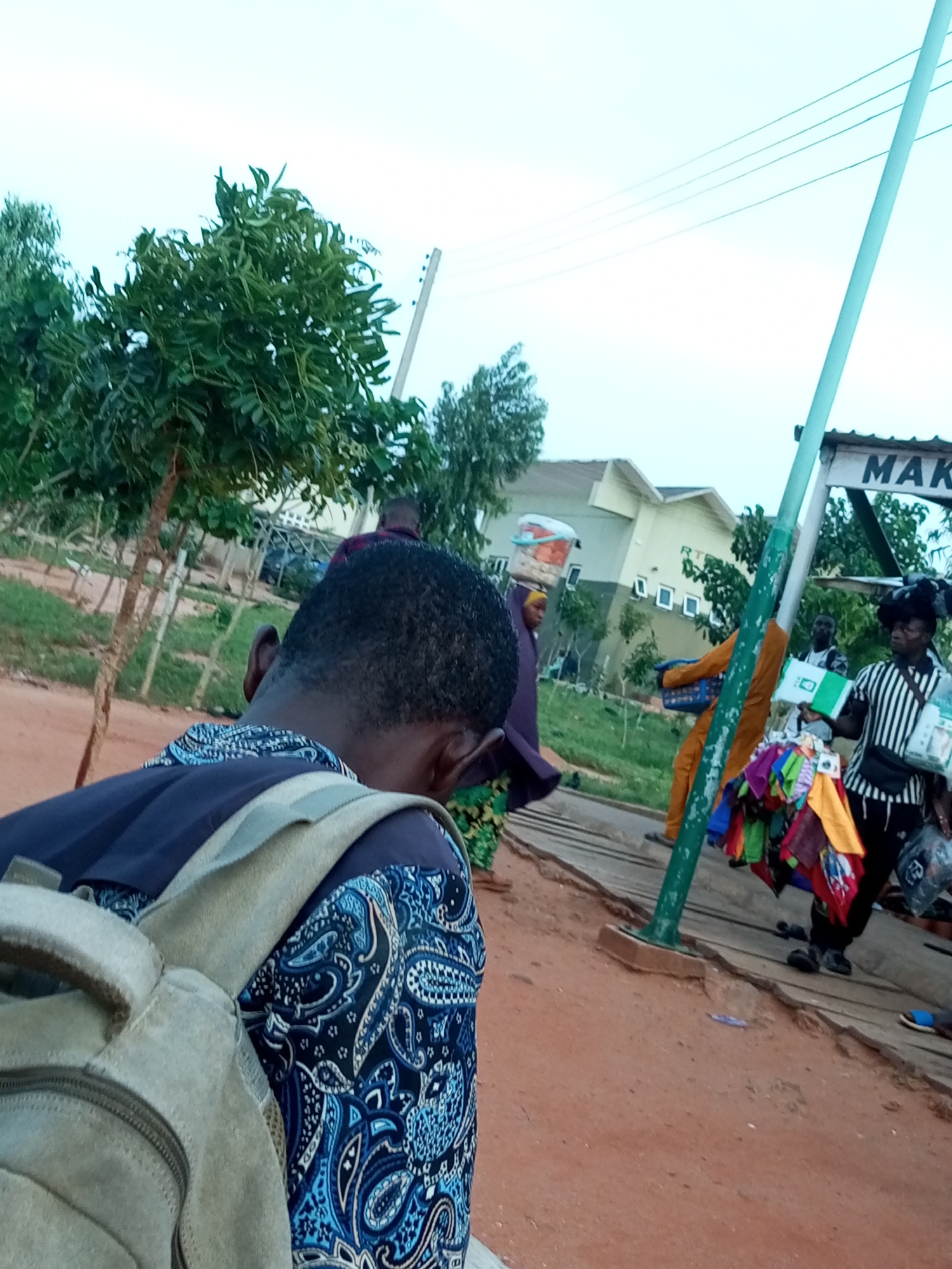 This media file depicts an outdoor scene in Gombe, Nigeria, likely in the late afternoon or early evening as indicated by the soft lighting. The setting appears to be a roadside market or busy walkway, with a sandy, reddish-brown ground and a wooden walkway or bridge in the mid-ground.
Several people are visible. In the foreground, the back of a young person wearing a dark blue shirt with a white and blue paisley pattern and a light brown backpack is captured. Their head is bowed. Further back, several individuals are engaged in commerce. One person, possibly a vendor, is standing near a green post and a sign that reads "MAK," holding up colorful folded fabrics or scarves for display. Another person in an orange outfit is walking with a blue basket. A woman is seen walking away from the camera with a bucket balanced on her head. Other figures are present in the background, suggesting a bustling environment.
The weather appears to be clear, with a bright, overcast sky. The scene conveys a sense of everyday life and commercial activity in a Nigerian town. The overall impression is one of a lively street scene with vendors and pedestrians.