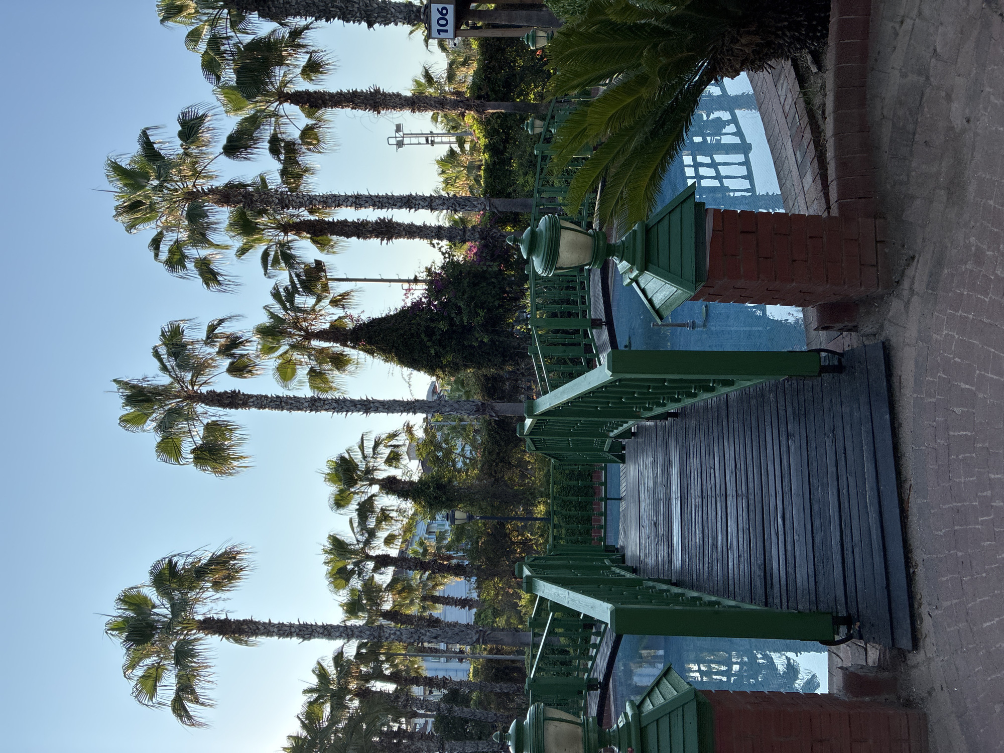 This image depicts a wooden bridge painted green, spanning a narrow body of blue water. The bridge has a weathered appearance, with visible planks and wooden railings. On either side of the bridge's entrance, there are brick pillars topped with green, pagoda-like structures housing lamps. Palm trees of varying heights dominate the background, with some reaching towards a clear blue sky. Green fences and lush vegetation are also present. In the distance, a building can be seen amongst the trees. A sign with the number "106" is visible to the right. The overall scene suggests an outdoor recreational area, possibly a resort or park, within the city of Alanya, Turkey.