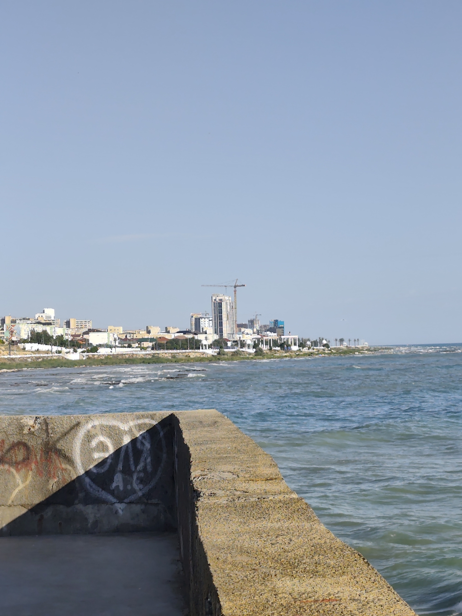 A bright, sunny day with a clear blue sky over a coastal city. In the foreground, a concrete structure, possibly a pier or breakwater, is visible with some graffiti on its side. The sea stretches out beyond, with visible waves and a slight chop, indicating a breezy day. In the middle ground, a city skyline is depicted, with a mix of modern high-rise buildings, some under construction with cranes visible, and lower-rise residential or commercial buildings. Palm trees can be seen scattered along the coastline, suggesting a warm climate. The overall scene conveys a sense of urban development along the coast. The time of day appears to be mid-morning or afternoon, given the bright sunlight and long shadows. The location is Aktau, Kazakhstan. No discernible emotions are conveyed.