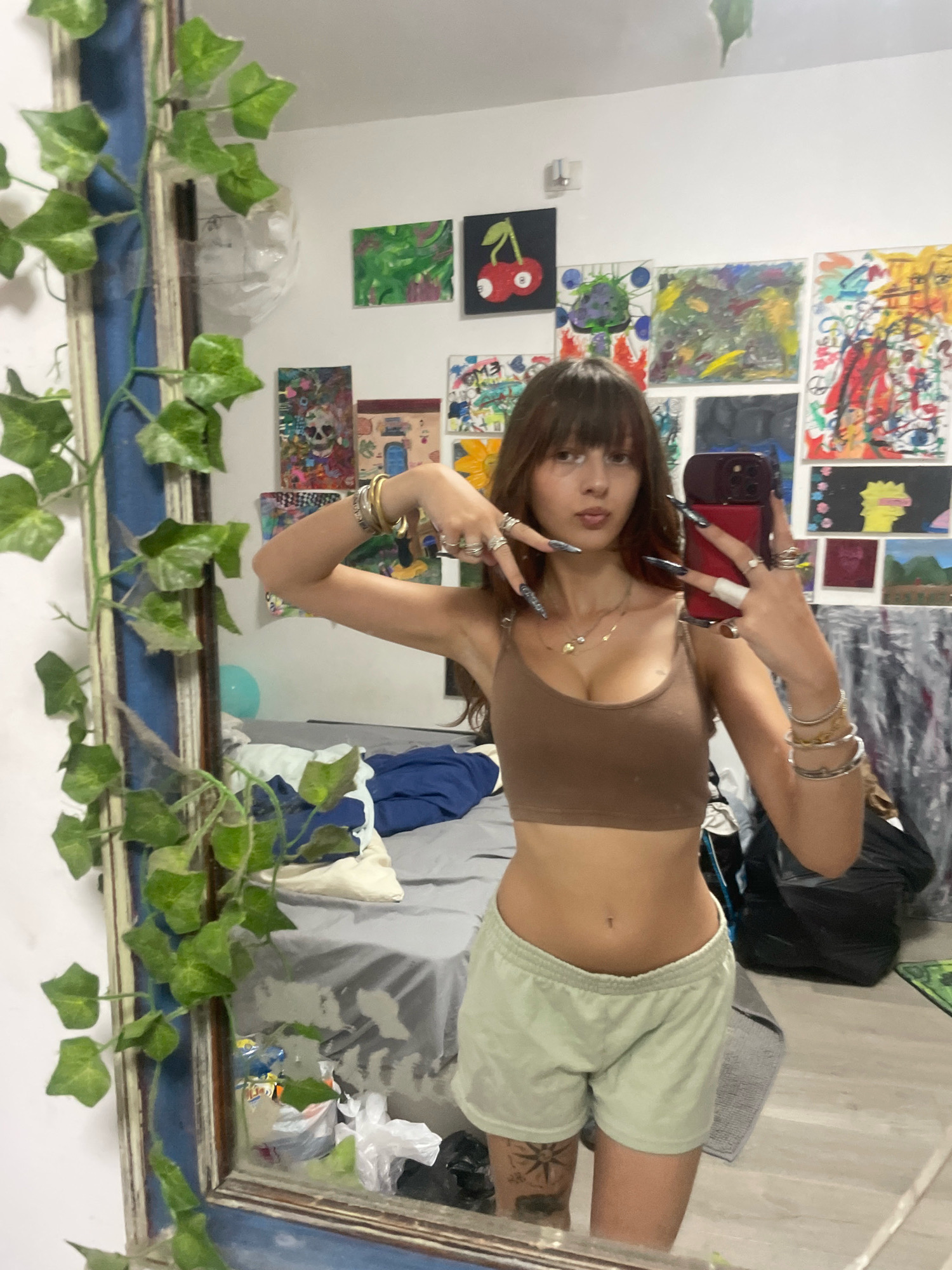 A young woman with long brown hair and bangs is taking a mirror selfie in what appears to be a bedroom. She is wearing a brown crop top and light green shorts. She has numerous rings on her fingers and bracelets on her wrists. The room is decorated with several pieces of art on the wall, and artificial ivy is draped around a large mirror to her left. The background shows a bed with gray sheets and some clothing. The overall impression is of a casual, lived-in space. Without any additional information, it is impossible to determine the specific location within the city of Raanana, Israel.