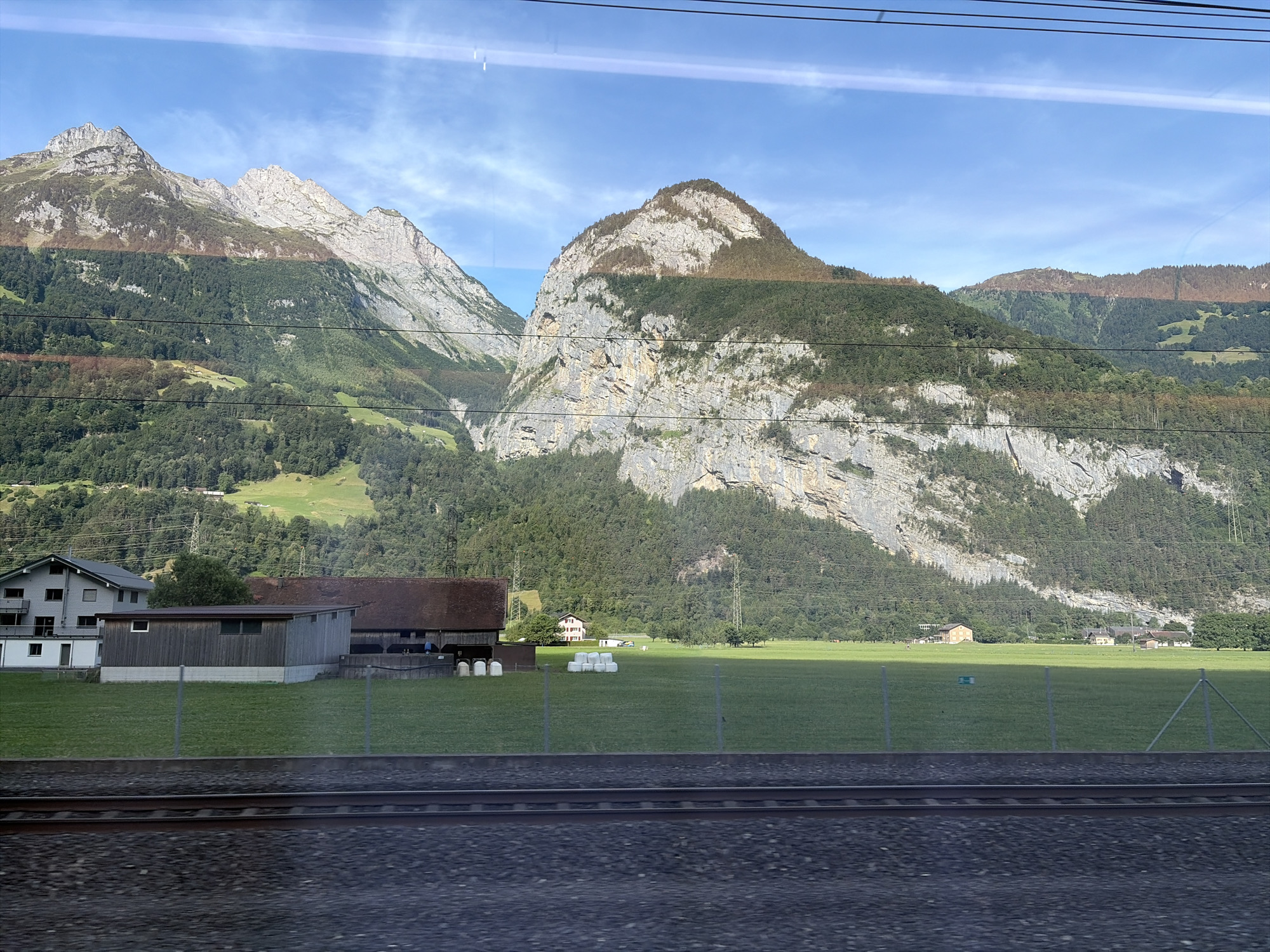 The image depicts a scenic view of the Swiss Alps, likely taken from a train moving parallel to a rural landscape. In the foreground, a section of train tracks with ballast is visible. Beyond the tracks, a bright green grassy field stretches towards a cluster of rural buildings. On the left, a modern gray residential building with balconies stands next to a long, dark wooden barn or storage structure. A row of white hay bales is neatly stacked in the field. In the middle ground, several farmhouses and other rural structures are scattered across the landscape, some with red roofs. The background is dominated by imposing, rocky mountains covered in dense green forests. The sky is a clear blue with a few wispy clouds. The image appears to be captured in the late afternoon or early evening, given the lighting and shadows. The location is identified as Schattdorf, Switzerland, which is consistent with the mountainous terrain and rural architecture.