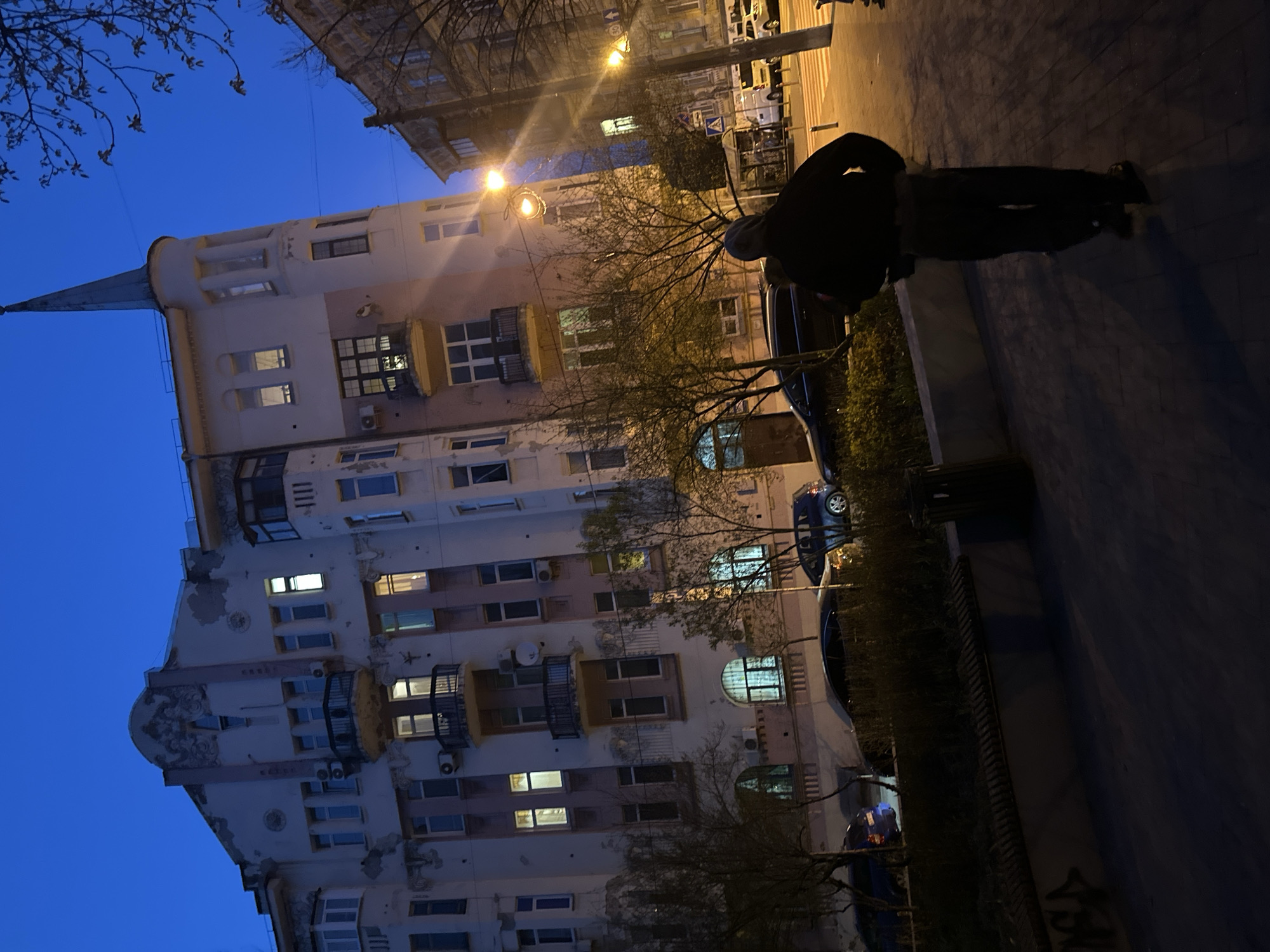 A street scene in Kyiv, Ukraine, at dusk. A tall, ornate building with a spire, multiple balconies, and illuminated windows is on the left. A person wearing a dark jacket and pants is standing on a sidewalk on the right side of the building. A street lamp illuminates the crosswalks. Several parked vehicles are observed. Bare trees are present in the area between the building and the street.